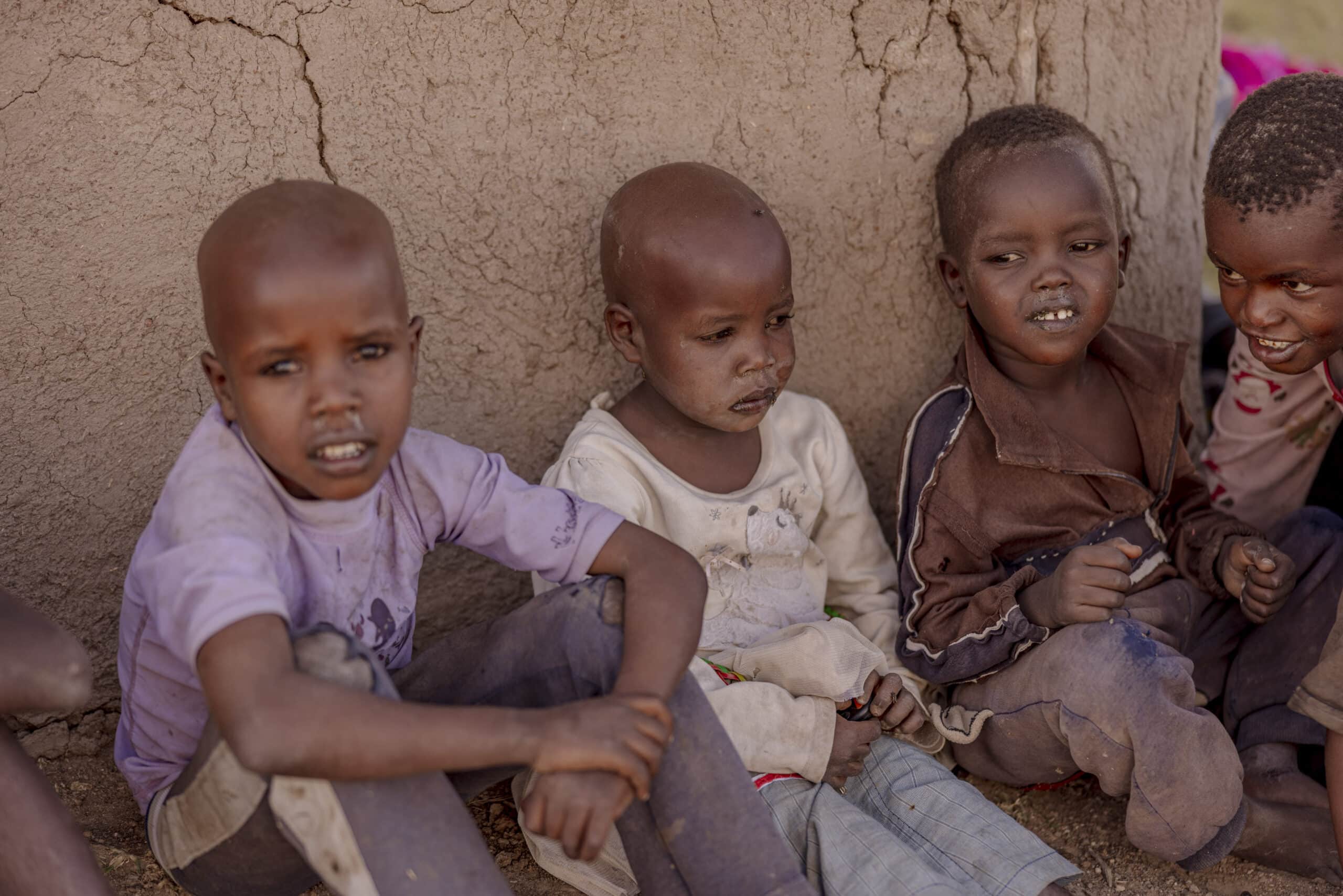 Vier kleine Kinder sitzen in einer ländlichen Umgebung zusammen an einer Lehmwand, sind verspielt und lächeln. Die Kinder scheinen zu spielen und die Gesellschaft des anderen zu genießen. © Fotografie Tomas Rodriguez