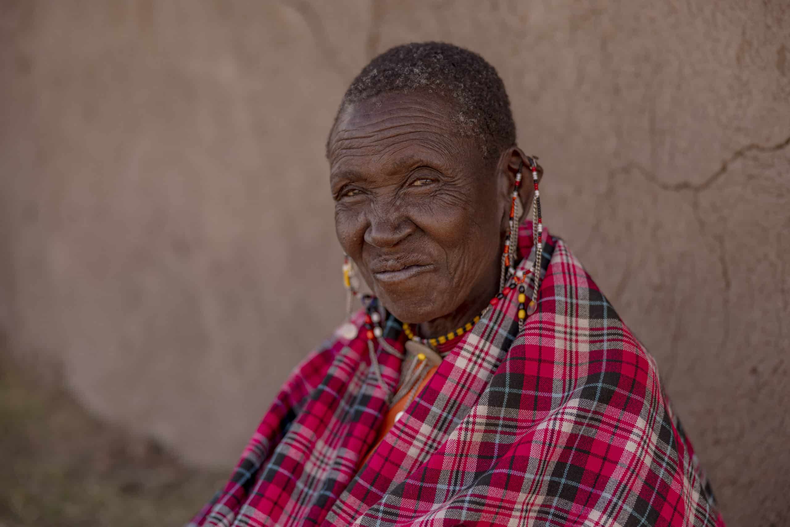 Eine ältere Frau, die traditionellen Massai-Perlenschmuck und eine rote Shuka trägt, lehnt an einer hellbraunen Lehmwand und blickt sanft und nachdenklich. © Fotografie Tomas Rodriguez