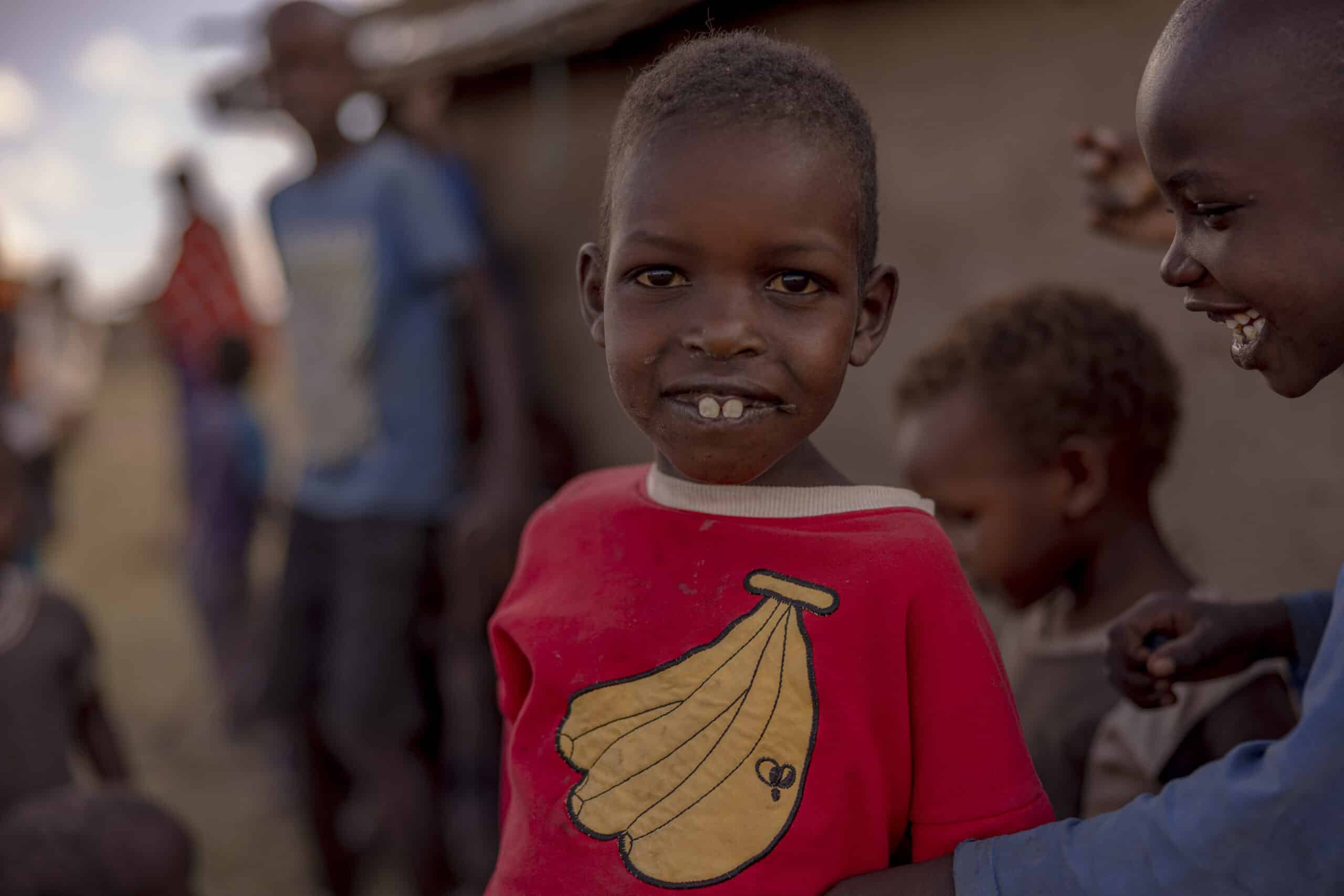 Ein kleiner Junge in einem roten Hemd mit Bananenaufdruck lächelt breit in die Kamera. Er steht zwischen anderen Kindern und einem Erwachsenen in einer ländlichen Umgebung. © Fotografie Tomas Rodriguez