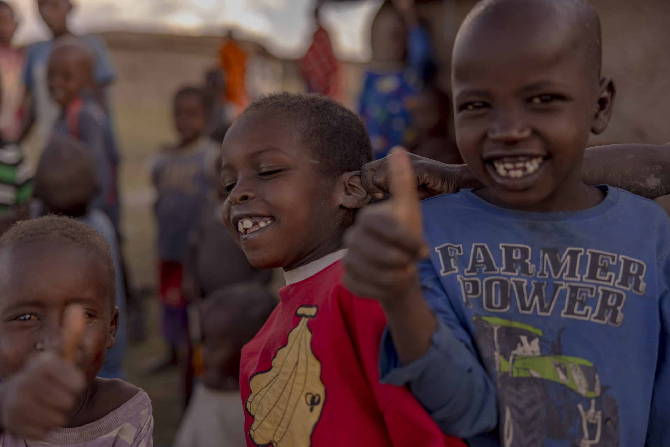 Zwei fröhliche afrikanische Kinder lächeln und zeigen in die Kamera, vor dem unscharfen Hintergrund sind andere Kinder in einer ländlichen Umgebung zu sehen. Ein Kind trägt ein rotes T-Shirt, das andere ein blaues „Farmer Power“-T-Shirt. © Fotografie Tomas Rodriguez
