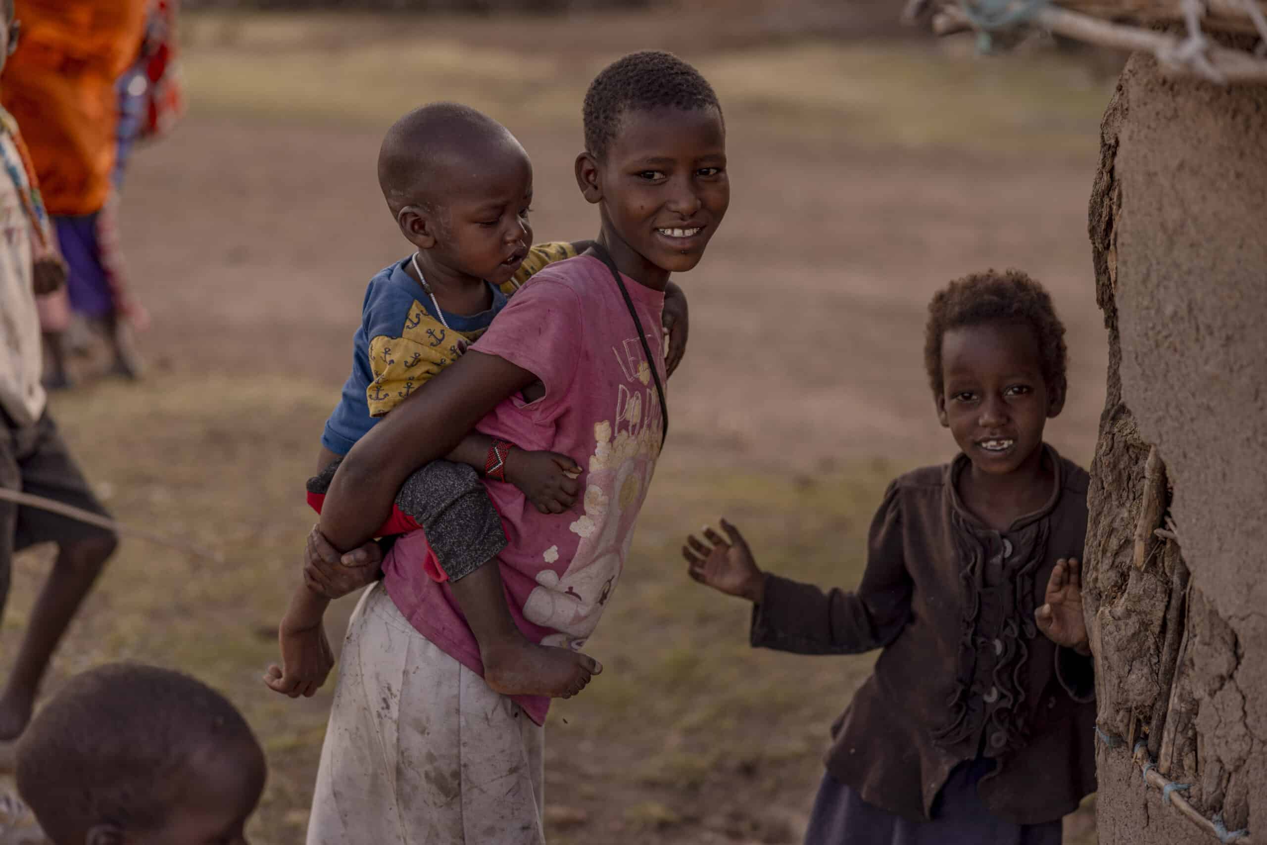 Ein junges Mädchen in ländlicher Umgebung trägt ein Kleinkind auf dem Rücken und lächelt in die Kamera, während ein anderes kleines Kind in der Nähe winkt. Sie sind umgeben von einer staubigen Landschaft und traditionellen Behausungen. © Fotografie Tomas Rodriguez