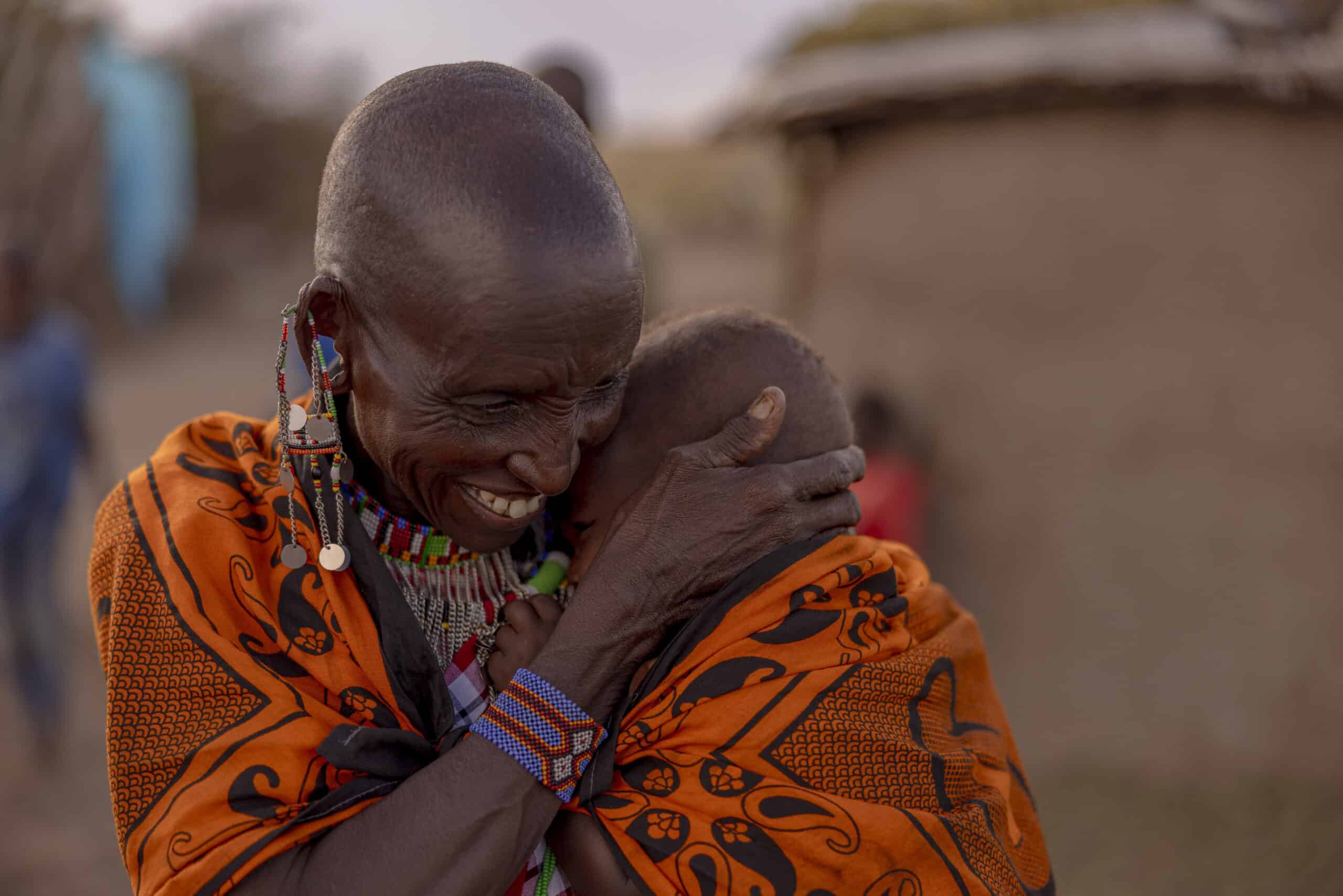 Eine ältere Frau in traditioneller Kleidung und mit Perlenketten umarmt in der Abenddämmerung freudig ein Kind in einer ländlichen Umgebung. © Fotografie Tomas Rodriguez