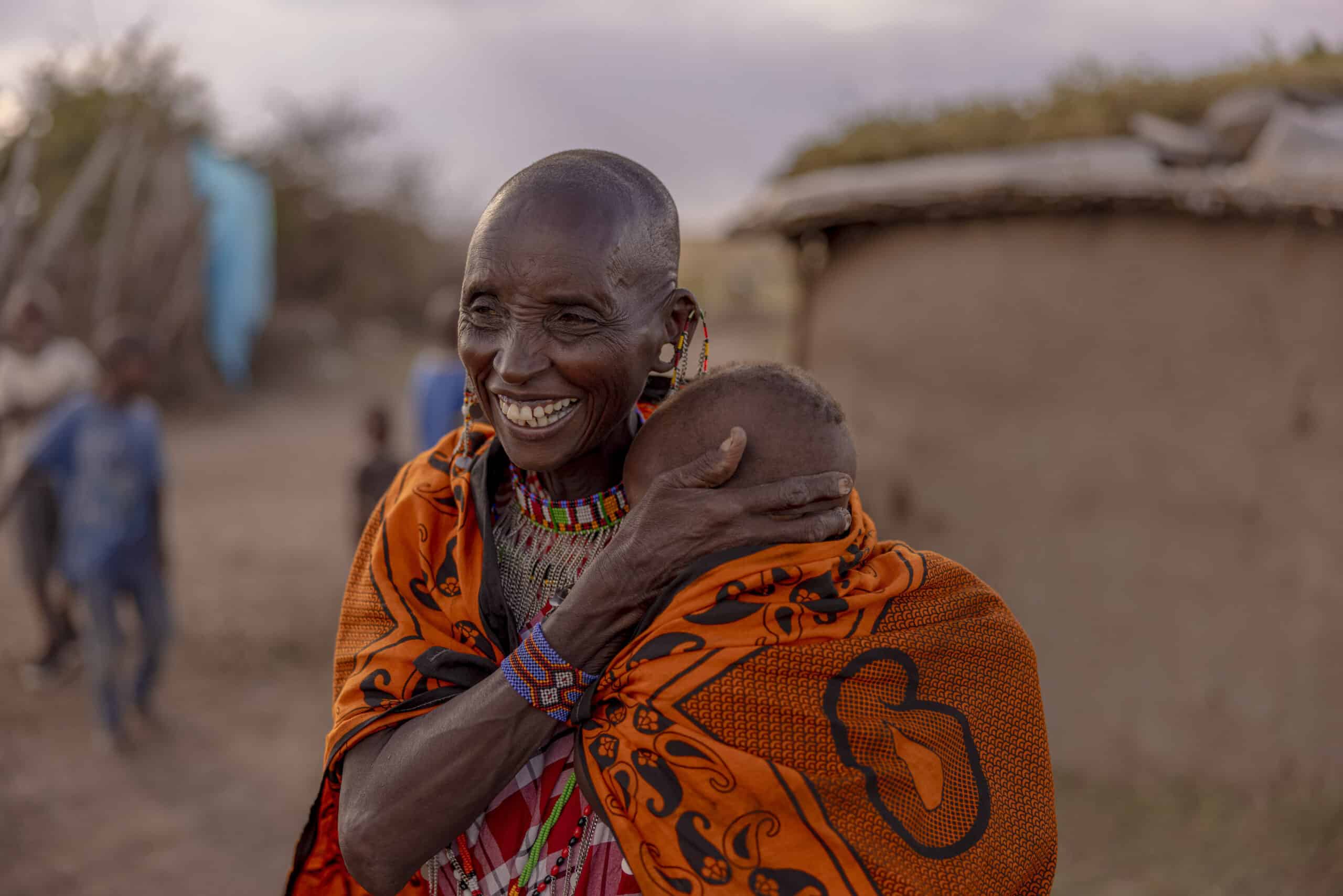 Eine ältere Frau in traditioneller Massai-Kleidung umarmt freudig ein Kind. Beide lachen in einer ländlichen Umgebung in der Nähe einer Strohhütte, im Hintergrund spielende Kinder. © Fotografie Tomas Rodriguez