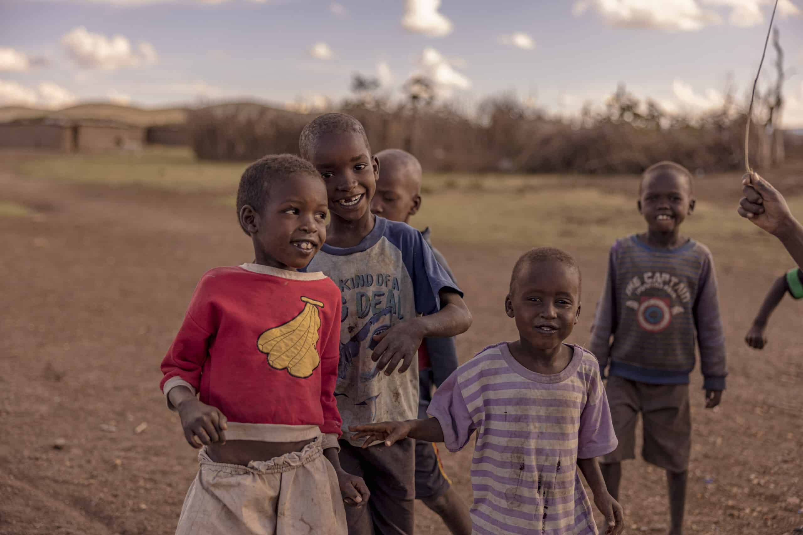 Vier kleine Kinder spielen auf einem Feld mit rustikalen Hütten im Hintergrund unter einem klaren Himmel. Sie lächeln und blicken in die Kamera. © Fotografie Tomas Rodriguez