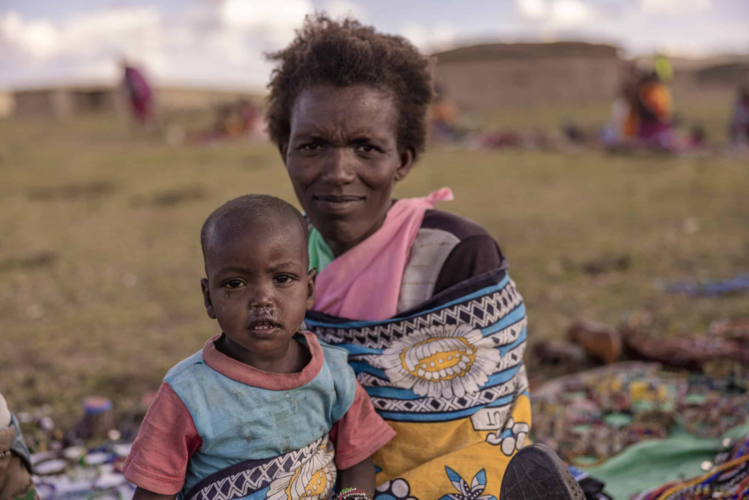 Eine Mutter in bunter Kleidung hält ihr kleines Kind in einer ländlichen Landschaft mit Hütten und Menschen im Hintergrund. Beide haben nachdenkliche Ausdrücke. © Fotografie Tomas Rodriguez