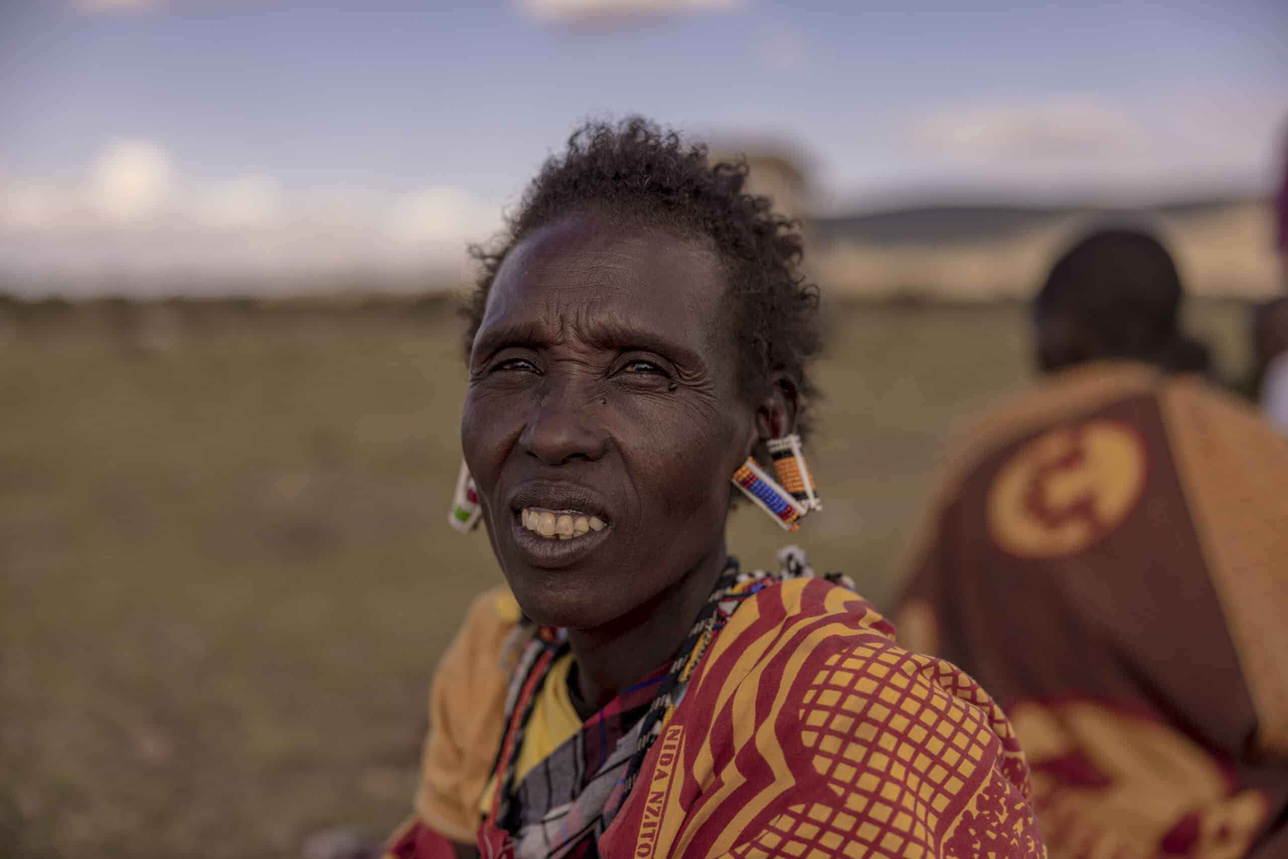 Eine Massai-Frau in traditioneller Kleidung, mit Perlenohrringen und einem leuchtend roten Shuka, lächelt im Freien vor dem unscharfen Hintergrund einer Wiese und einer weiteren Person in ähnlicher Kleidung. © Fotografie Tomas Rodriguez