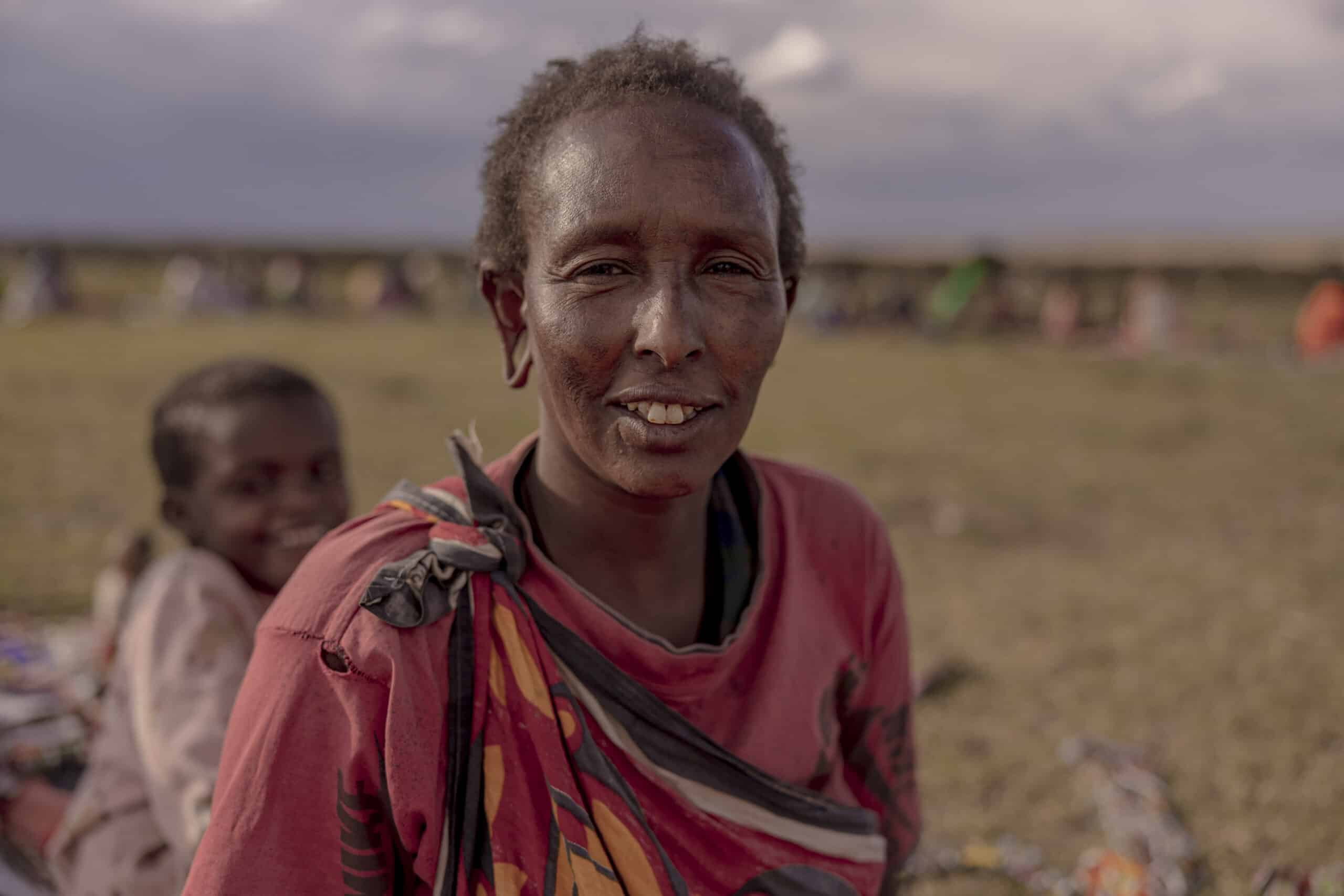 Ein Porträt einer lächelnden Massai-Frau in traditioneller Kleidung vor dem unscharfen Hintergrund einer Wiese und sitzender Menschen. Ihr Gesichtsausdruck ist warmherzig und die Szene spielt im Freien bei Tageslicht. © Fotografie Tomas Rodriguez