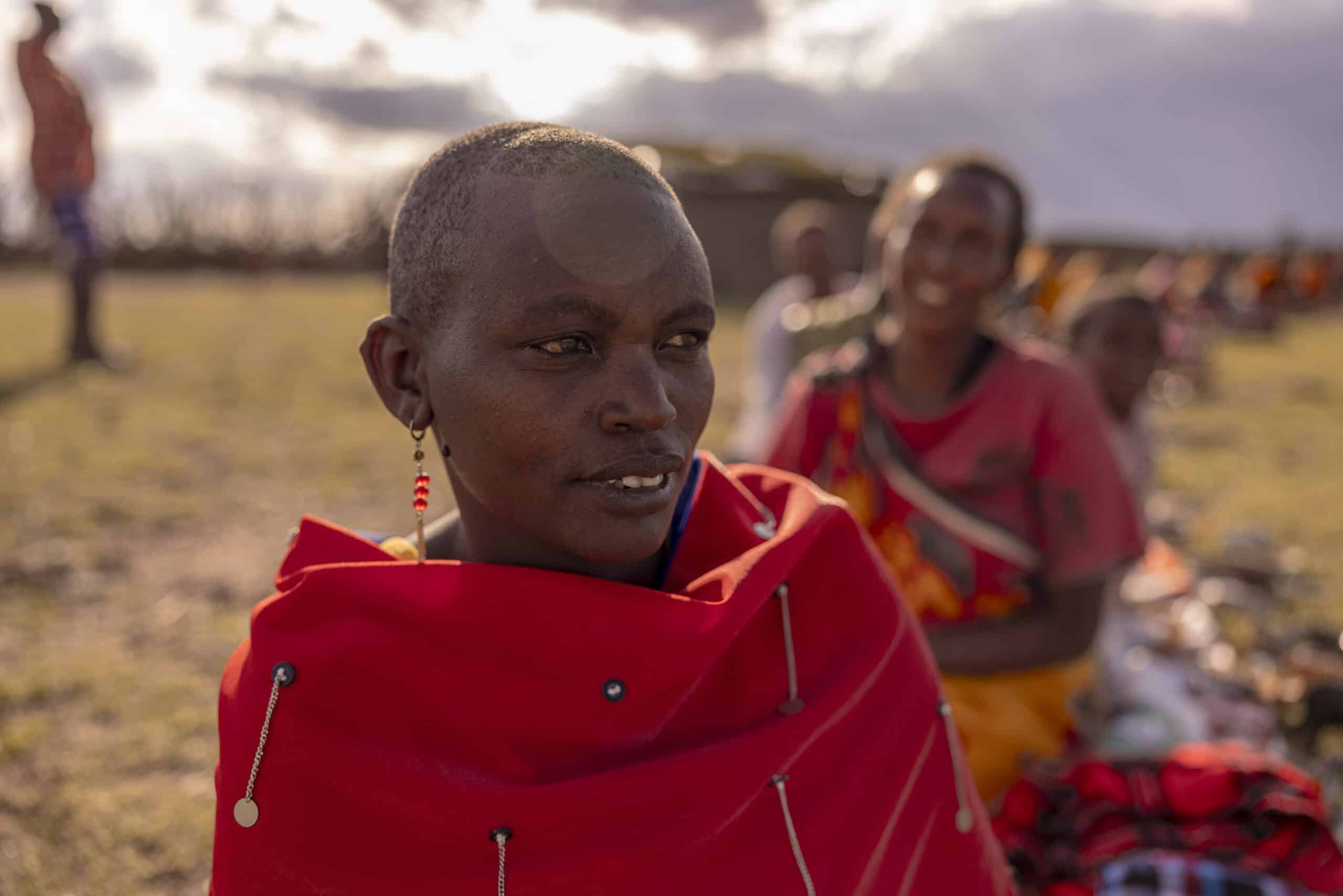 Eine Massai-Frau im traditionellen roten Shuka, mit dekorativen Ohrringen und rasiertem Kopf blickt in einer Außenumgebung nachdenklich zur Seite. Im Hintergrund sind andere Massai zu sehen. © Fotografie Tomas Rodriguez