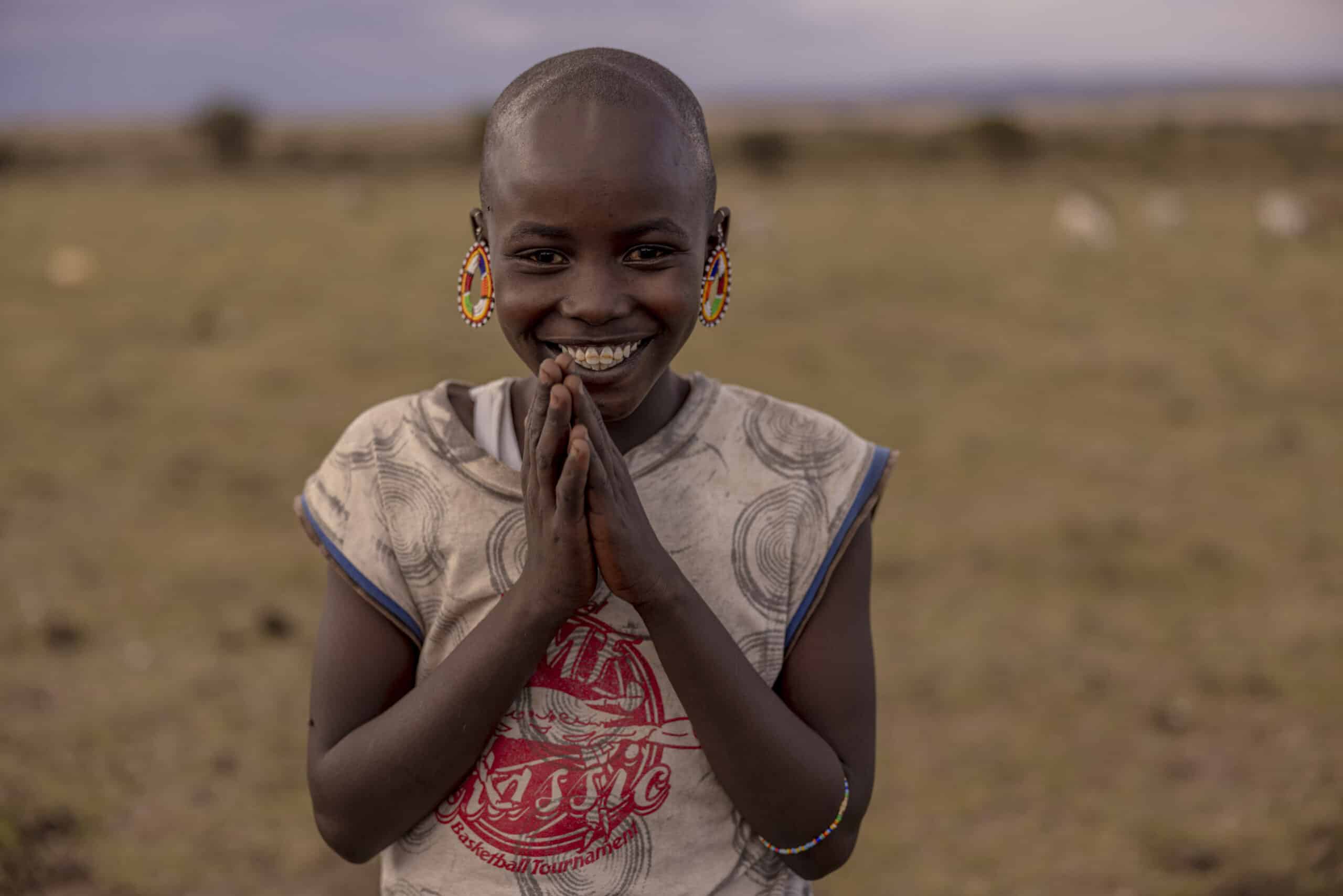 Ein fröhliches junges Mädchen mit rasiertem Kopf und großen Perlenohrringen lächelt breit und hat die Hände gefaltet. Es steht auf einer Wiese unter einem bewölkten Himmel. © Fotografie Tomas Rodriguez