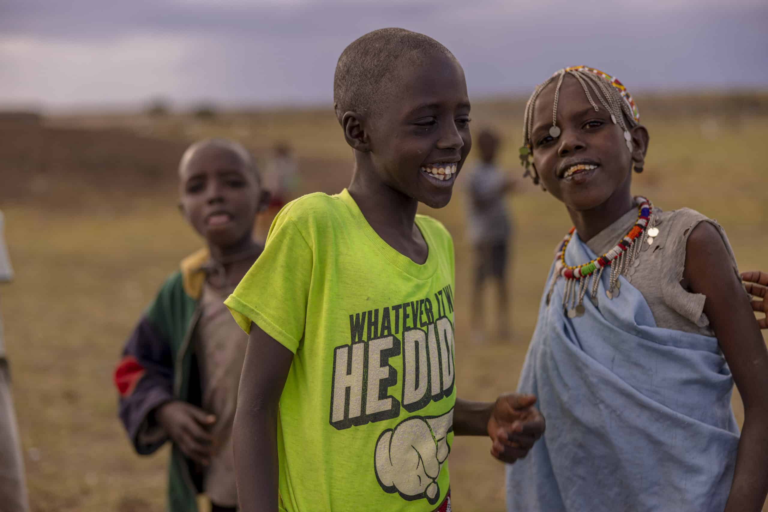 Drei afrikanische Kinder auf einem Feld in der Abenddämmerung; ein Mädchen mit einer Perlenkette und ein Junge in einem hellgrünen Hemd lachen zusammen, ein weiterer Junge ist im Hintergrund. © Fotografie Tomas Rodriguez