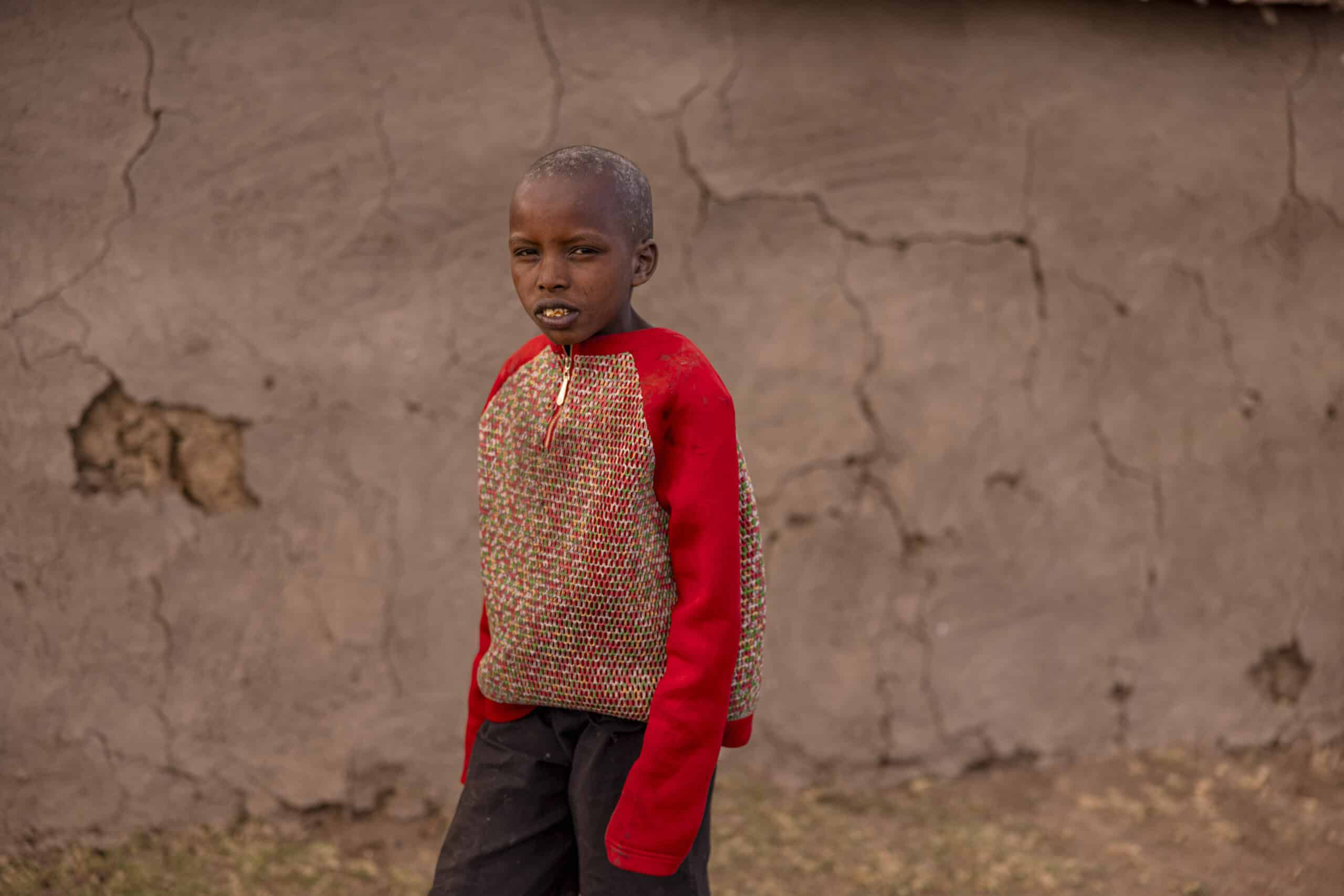 Ein kleiner Junge steht vor einer Wand mit großen Rissen. Er trägt einen roten Pullover und eine bunte Weste. Der Hintergrund ist unscharf, sodass die Aufmerksamkeit auf seinen nachdenklichen Gesichtsausdruck fällt. © Fotografie Tomas Rodriguez