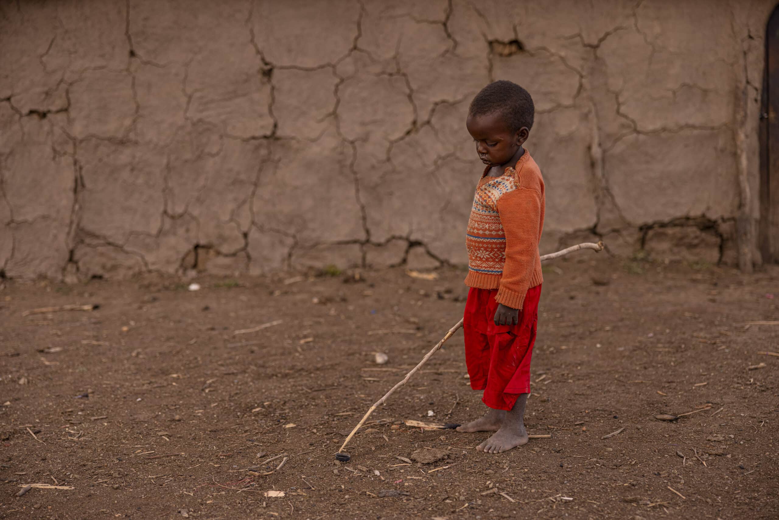 Ein kleines Kind steht barfuß auf einem Erdboden und spielt mit einem Stock und einem Reifen neben einer rissigen Lehmwand. Das Kind trägt einen rot-orangefarbenen Pullover und eine rote Hose und blickt nachdenklich nach unten. © Fotografie Tomas Rodriguez