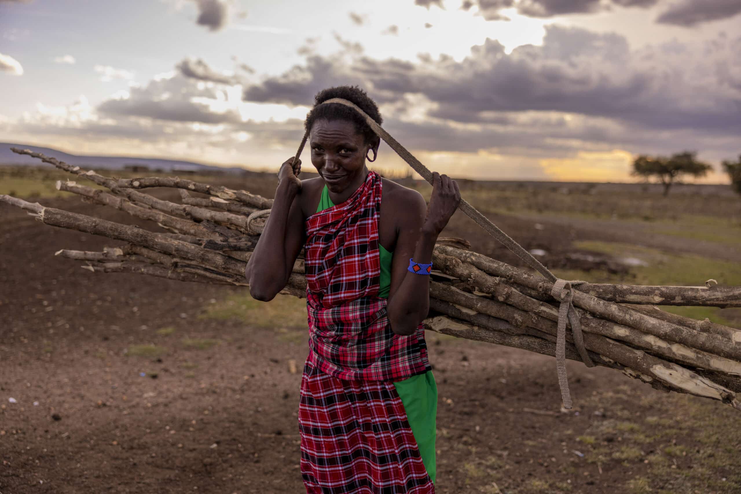 Eine Frau in traditioneller Massai-Kleidung trägt ein Bündel Stöcke auf dem Rücken in einer weiten, offenen Landschaft in der Abenddämmerung, über der sich dramatische Wolken auftürmen. © Fotografie Tomas Rodriguez