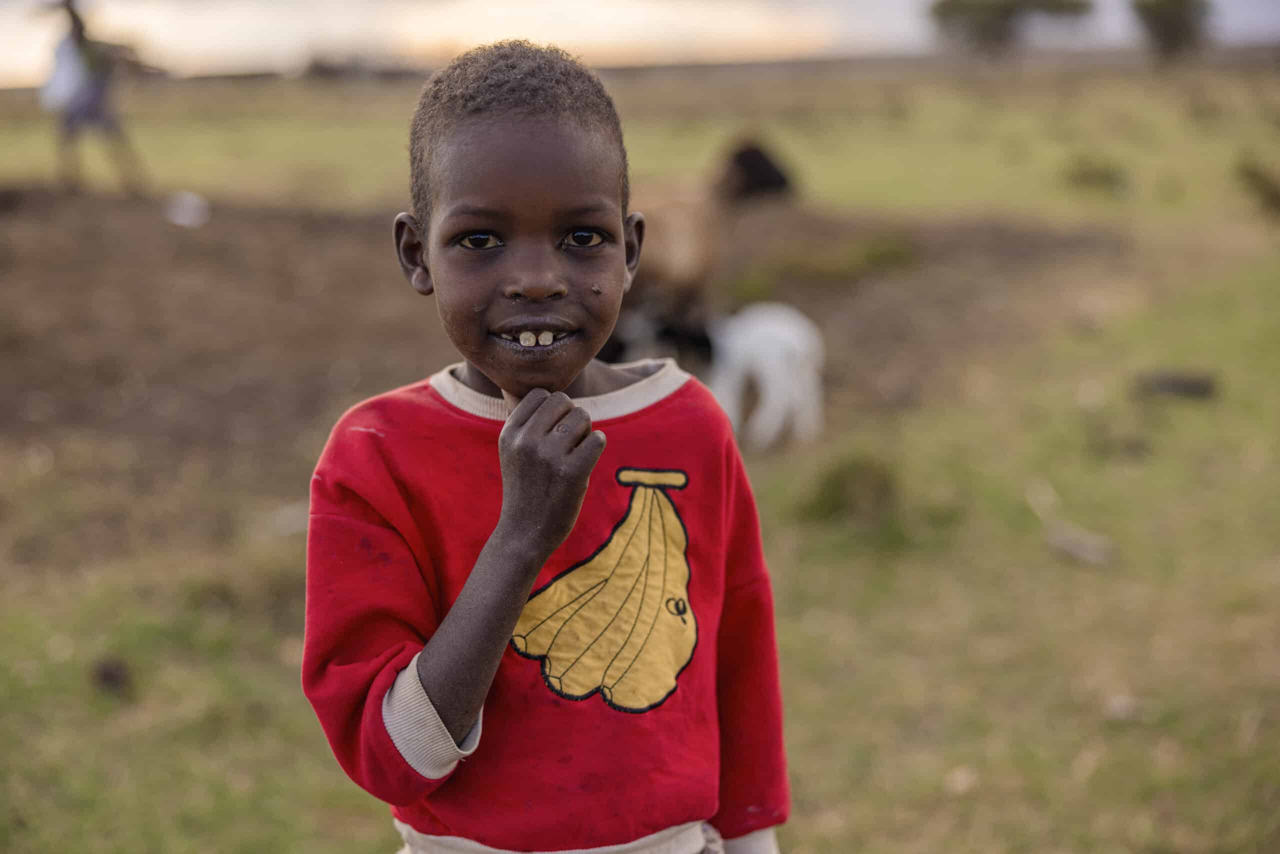 Ein kleines Kind mit einem strahlenden Lächeln und einem roten Pullover steht im Freien vor einer malerischen Kulisse mit schwachen Umrissen von Bäumen und Tieren. © Fotografie Tomas Rodriguez