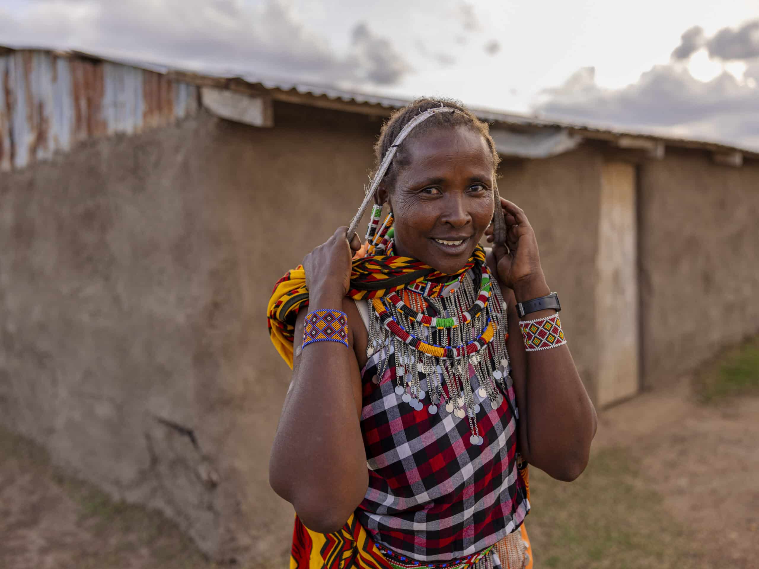 Eine lächelnde Frau in traditioneller kenianischer Kleidung, geschmückt mit leuchtenden Perlen und farbenfrohen Kleidungsstücken, steht vor rustikalen Gebäuden und rückt unter einem bewölkten Himmel ihre Halskette zurecht. © Fotografie Tomas Rodriguez