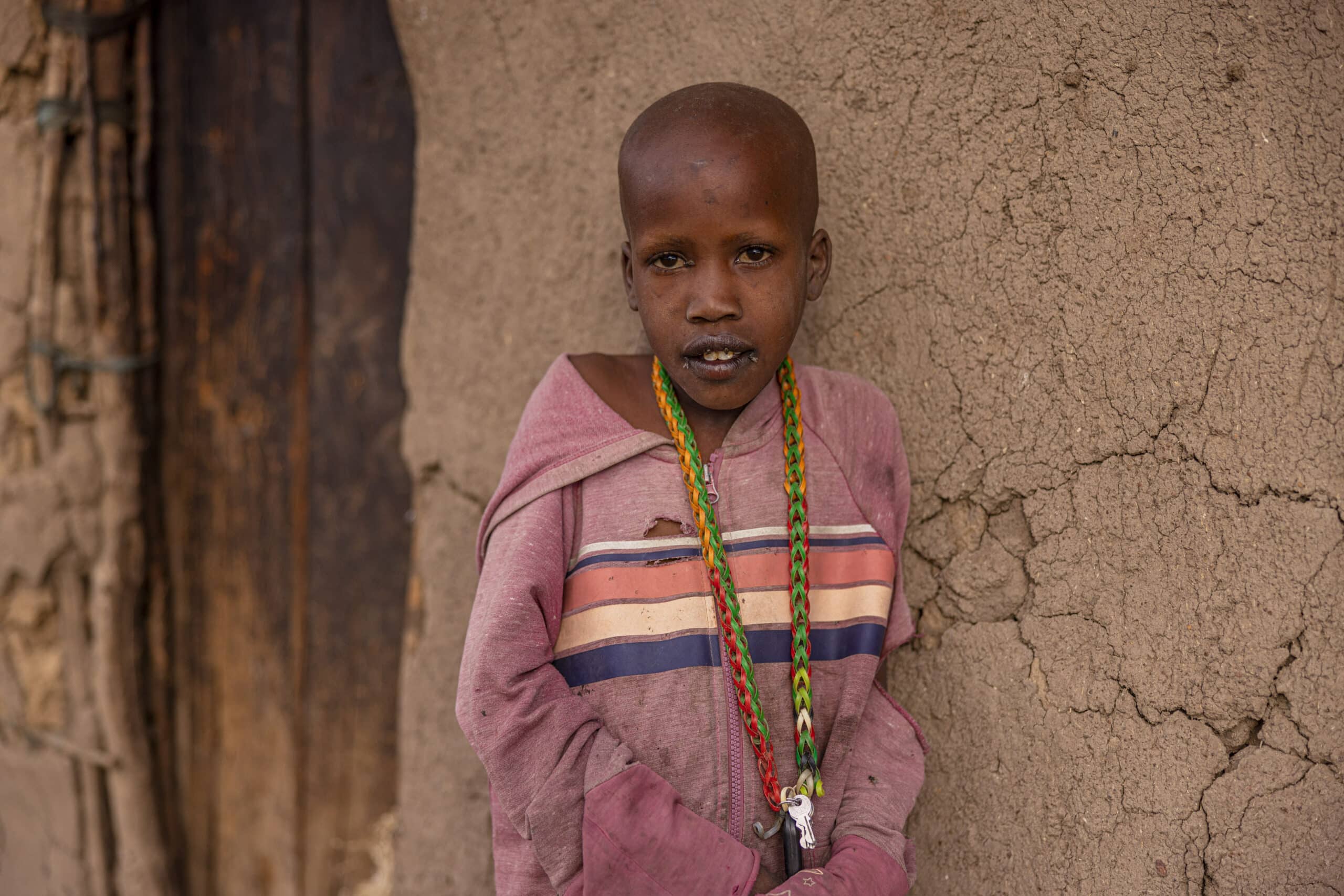Ein kleiner Junge mit rasiertem Kopf steht vor einer rissigen Lehmwand. Er trägt einen gestreiften Kapuzenpullover und bunte Perlenketten und blickt direkt in die Kamera. © Fotografie Tomas Rodriguez