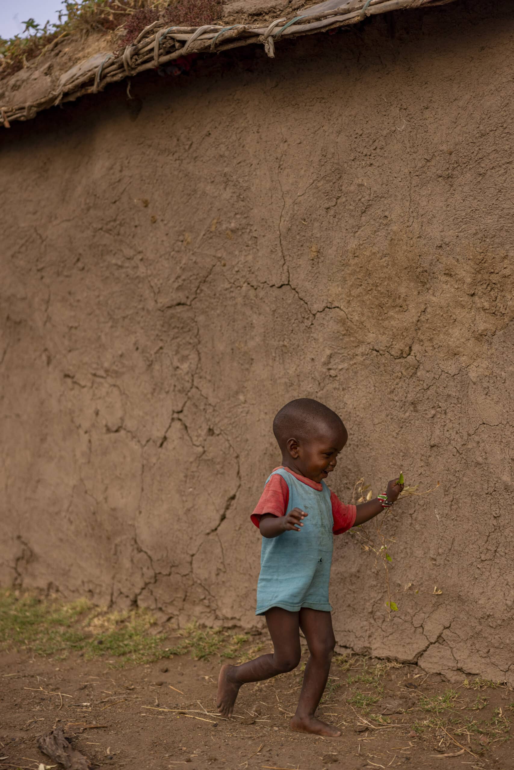 Ein kleines Kind in einem rot-blauen Outfit geht in einer ländlichen Umgebung an einer großen Lehmmauer mit Strohdach vorbei und hält einen kleinen Ast. © Fotografie Tomas Rodriguez