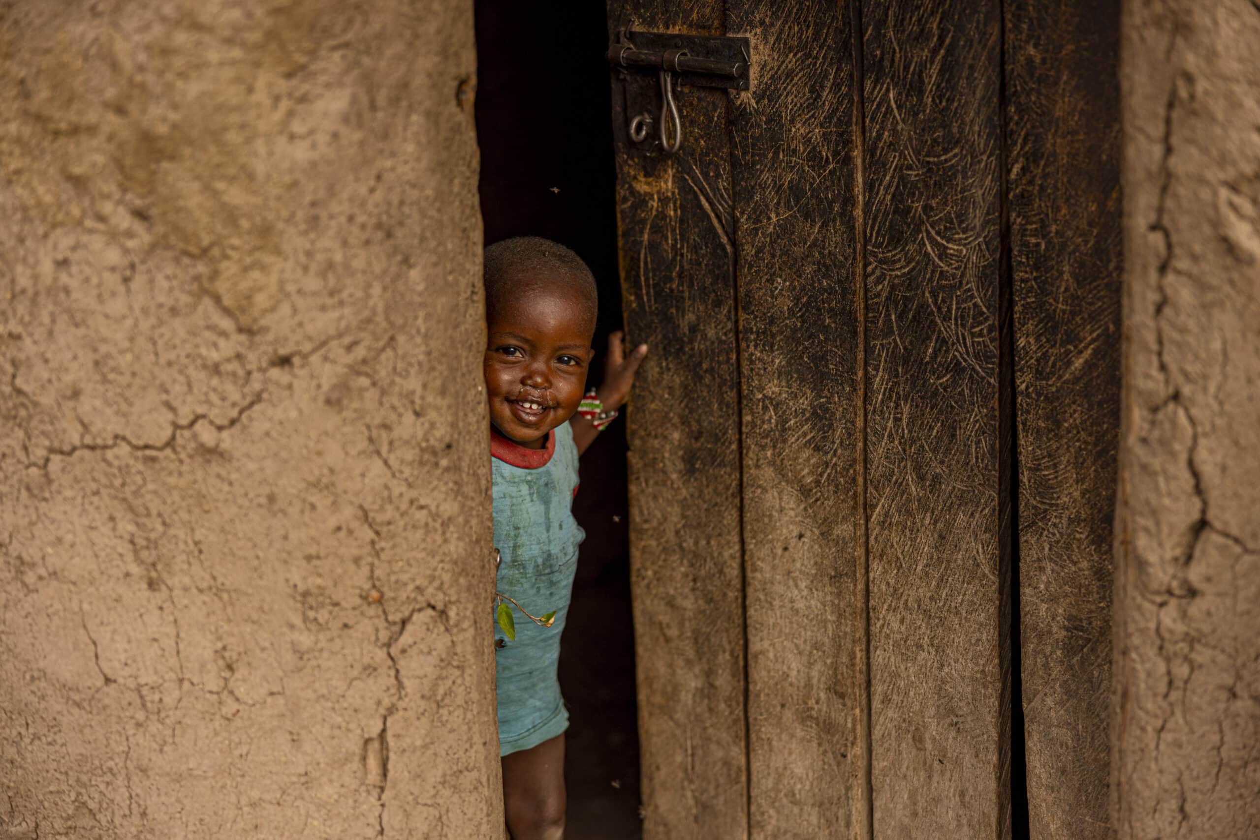 Ein kleines Kind lächelt, als es durch eine leicht geöffnete Holztür späht und den Blick auf eine rustikale Lehmwand und traditionelle Radierungen auf der Tür freigibt. © Fotografie Tomas Rodriguez