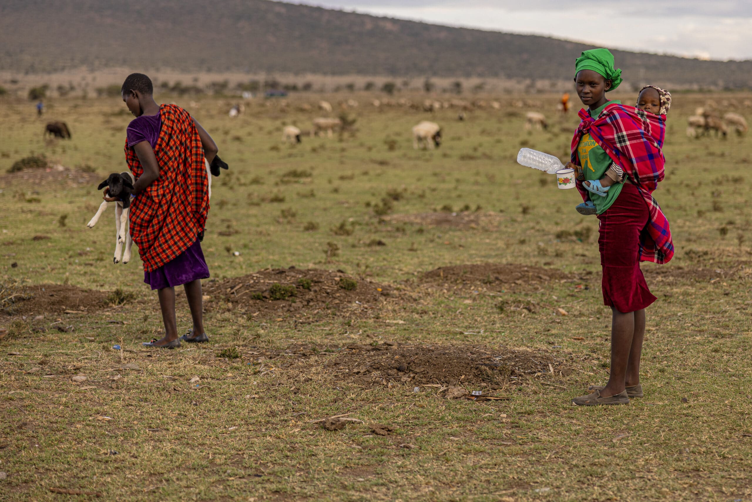 Zwei Massai-Frauen in traditioneller Kleidung. Eine hält ein Baby und einen Plastikbehälter, die andere blickt weg. In der Ferne streifen Ziegen umher. © Fotografie Tomas Rodriguez