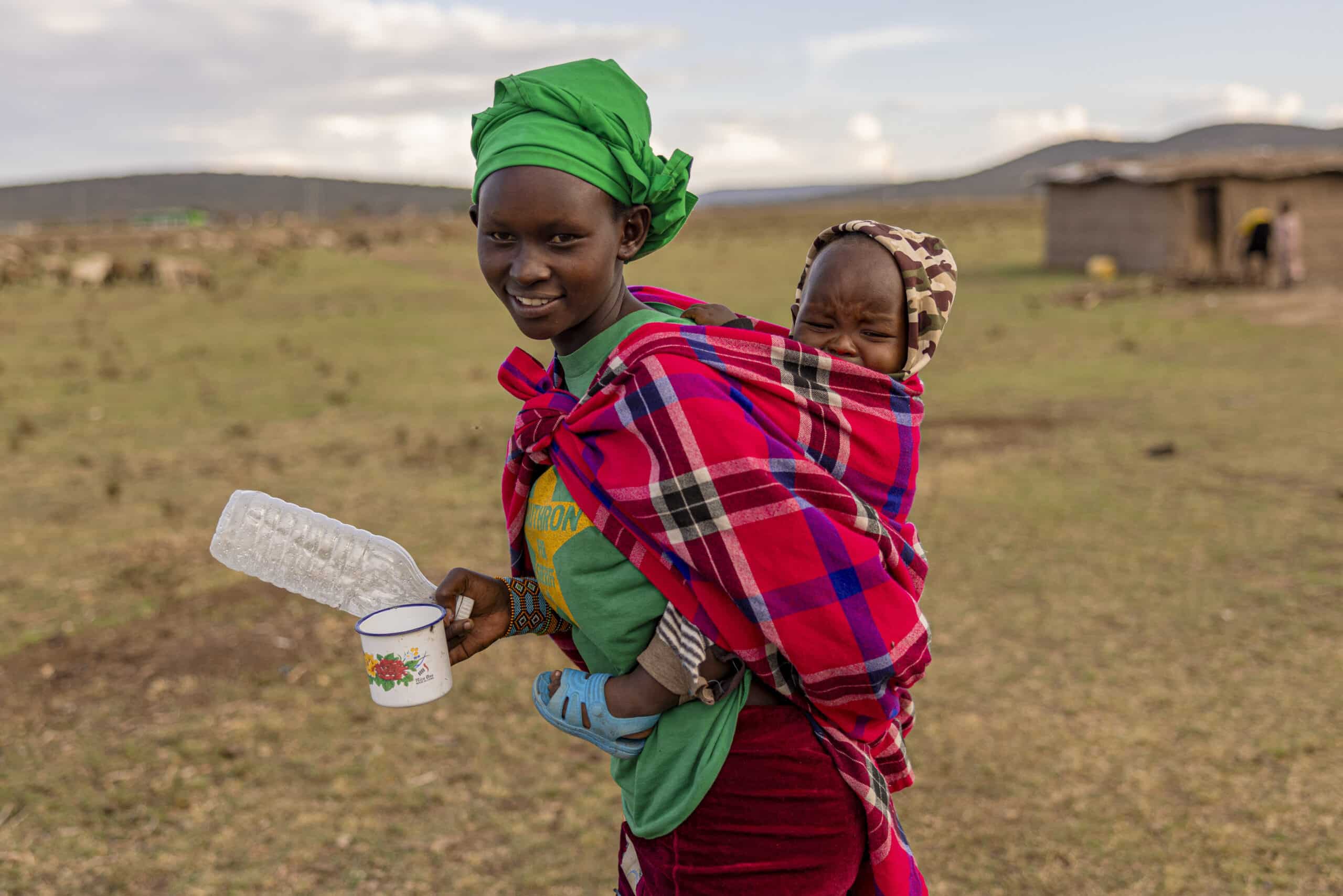 Eine junge Frau mit einem leuchtend grünen Kopftuch und einem karierten Schal lächelt, trägt ein Baby auf dem Rücken und hält eine leere Plastikflasche in der Hand, im Hintergrund sind eine ländliche Landschaft und Hütten zu sehen. © Fotografie Tomas Rodriguez
