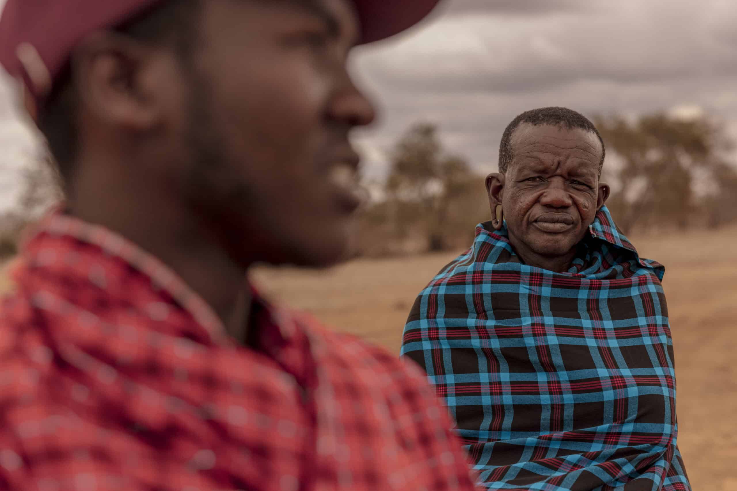 Zwei Männer in traditionellen Massai-Shukas, einer unscharf im Vordergrund, der andere scharf im Hintergrund, stehen in einer Savannenlandschaft. © Fotografie Tomas Rodriguez