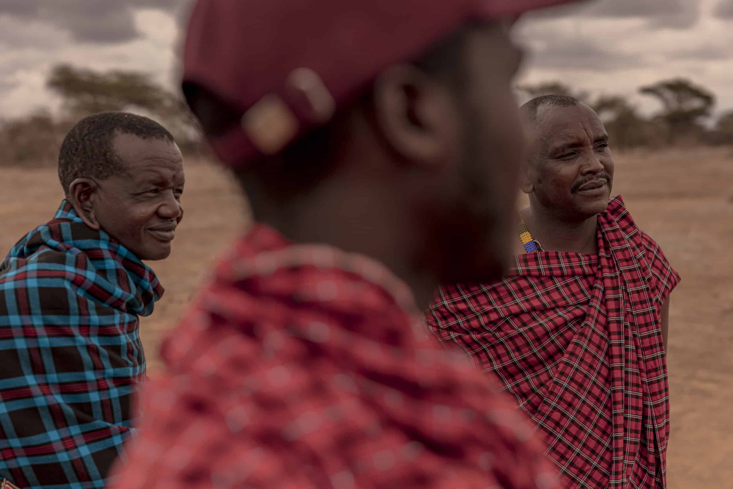 Drei Männer in traditionellen Massai-Shukas stehen im Freien vor einem bewölkten Himmel und der Savanne im Hintergrund. Zwei Männer blicken nachdenklich in die Ferne, während der dritte, verschwommen im Vordergrund, ihnen gegenübersteht. © Fotografie Tomas Rodriguez