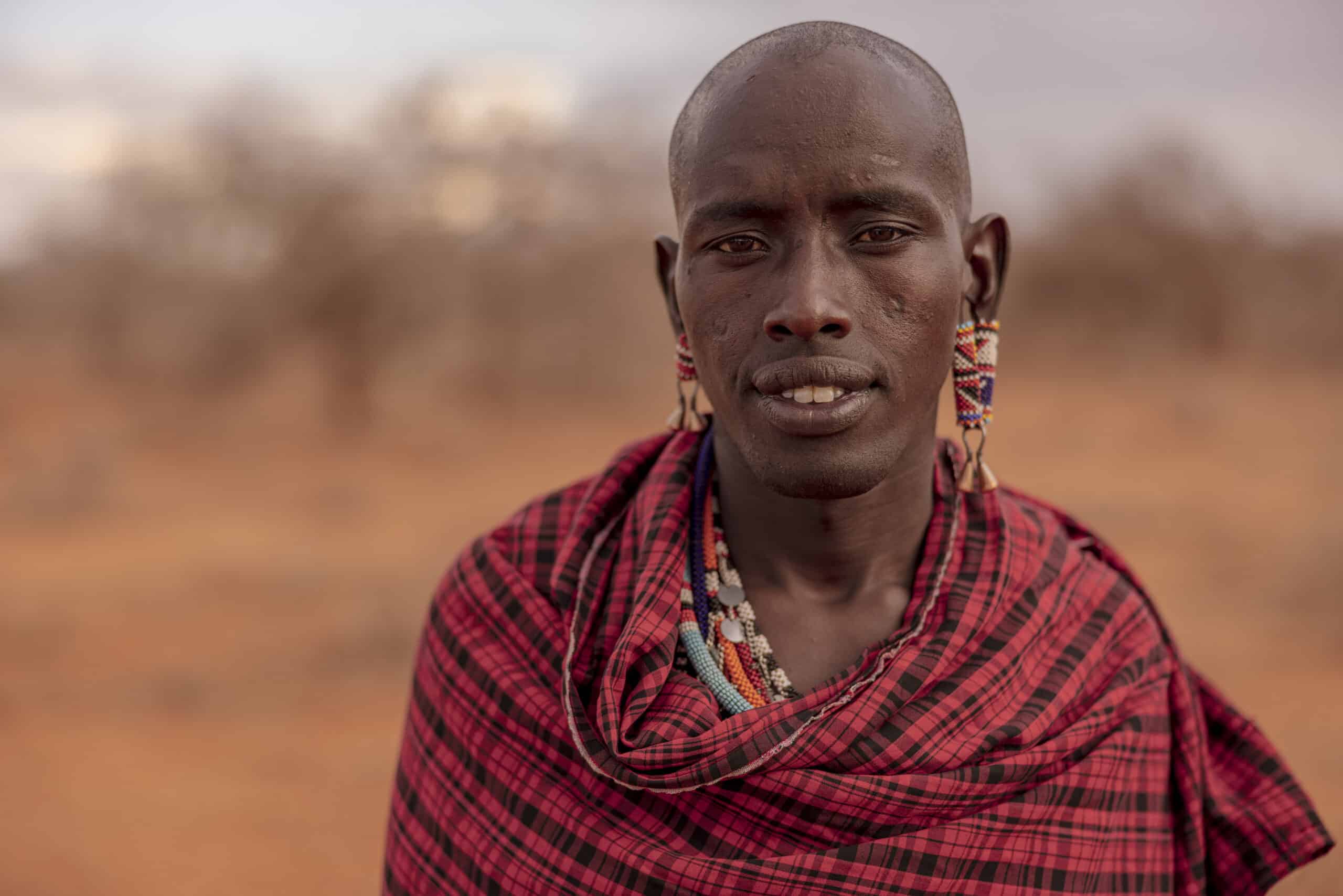 Ein Porträt eines Massai-Mannes in traditioneller Kleidung und Perlenstickerei, der vor einer verschwommenen, trockenen Landschaft steht. © Fotografie Tomas Rodriguez