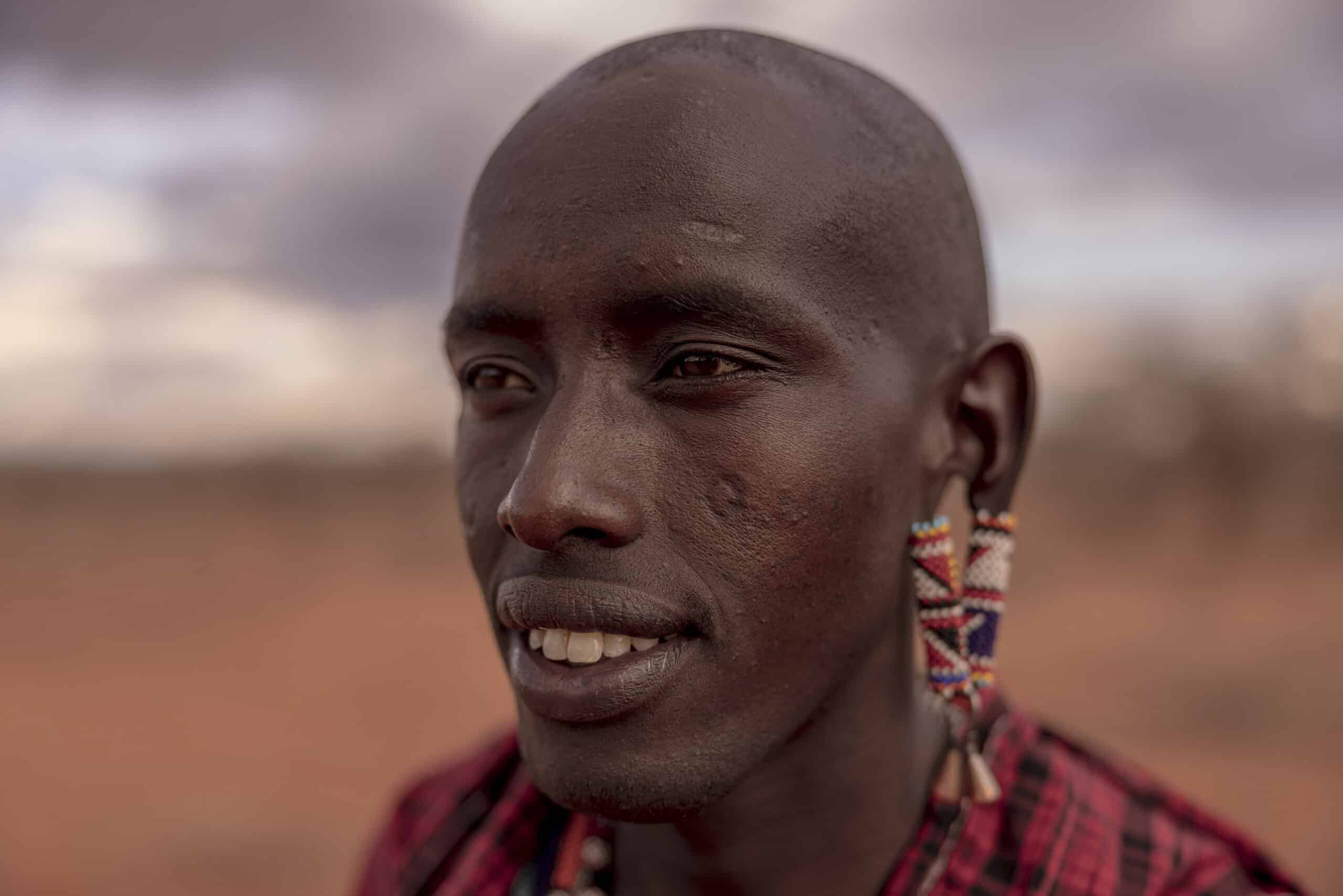 Nahaufnahme eines Massai-Mannes mit traditionellem Schmuck, darunter Perlenohrringe und eine rote Shuka, vor dem unscharfen Hintergrund einer halbtrockenen Landschaft. © Fotografie Tomas Rodriguez