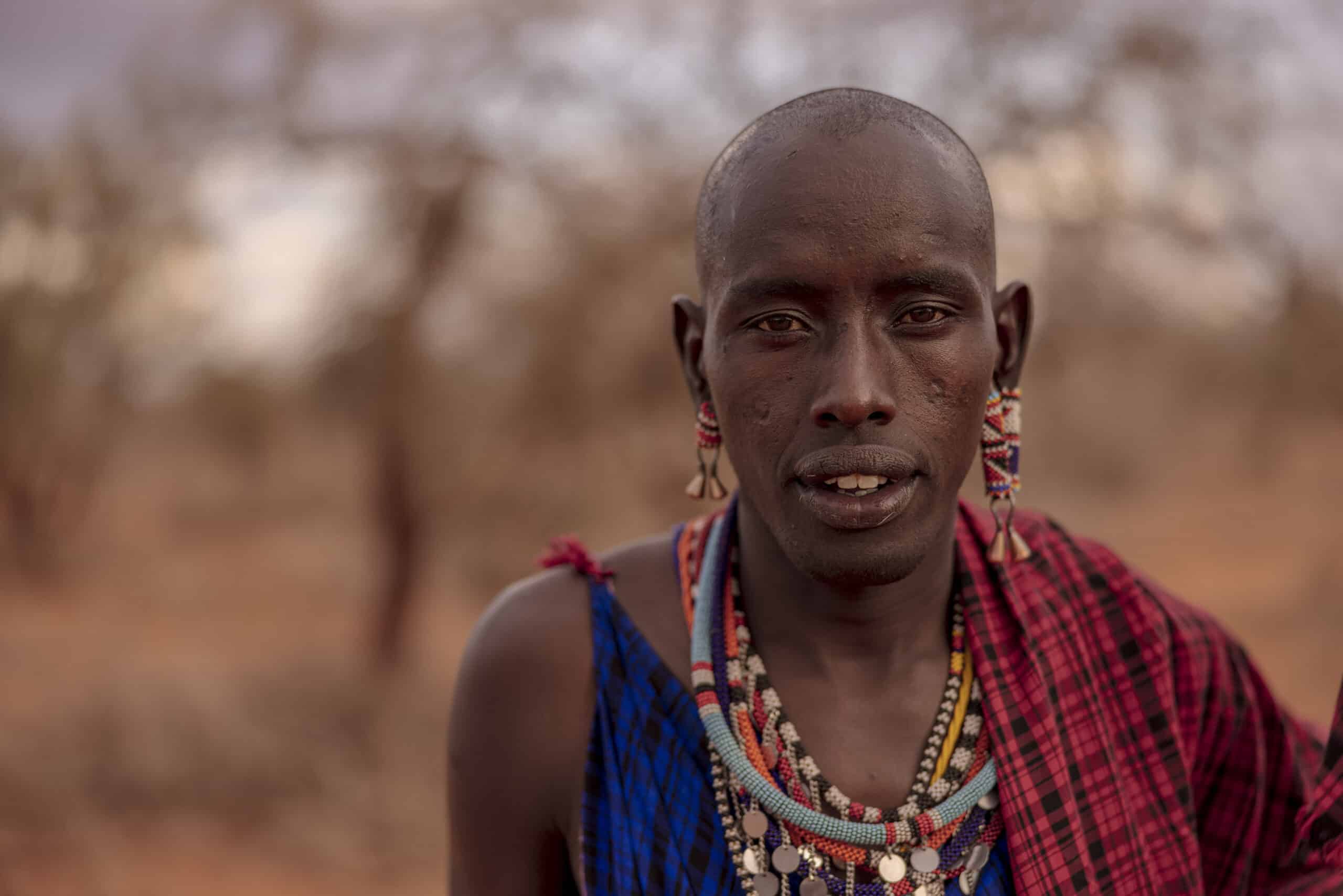 Ein Porträt eines Massai-Mannes mit traditionellem Schmuck und traditioneller Kleidung und heiterem Gesichtsausdruck, der in der Abenddämmerung in einer kargen Landschaft steht. © Fotografie Tomas Rodriguez