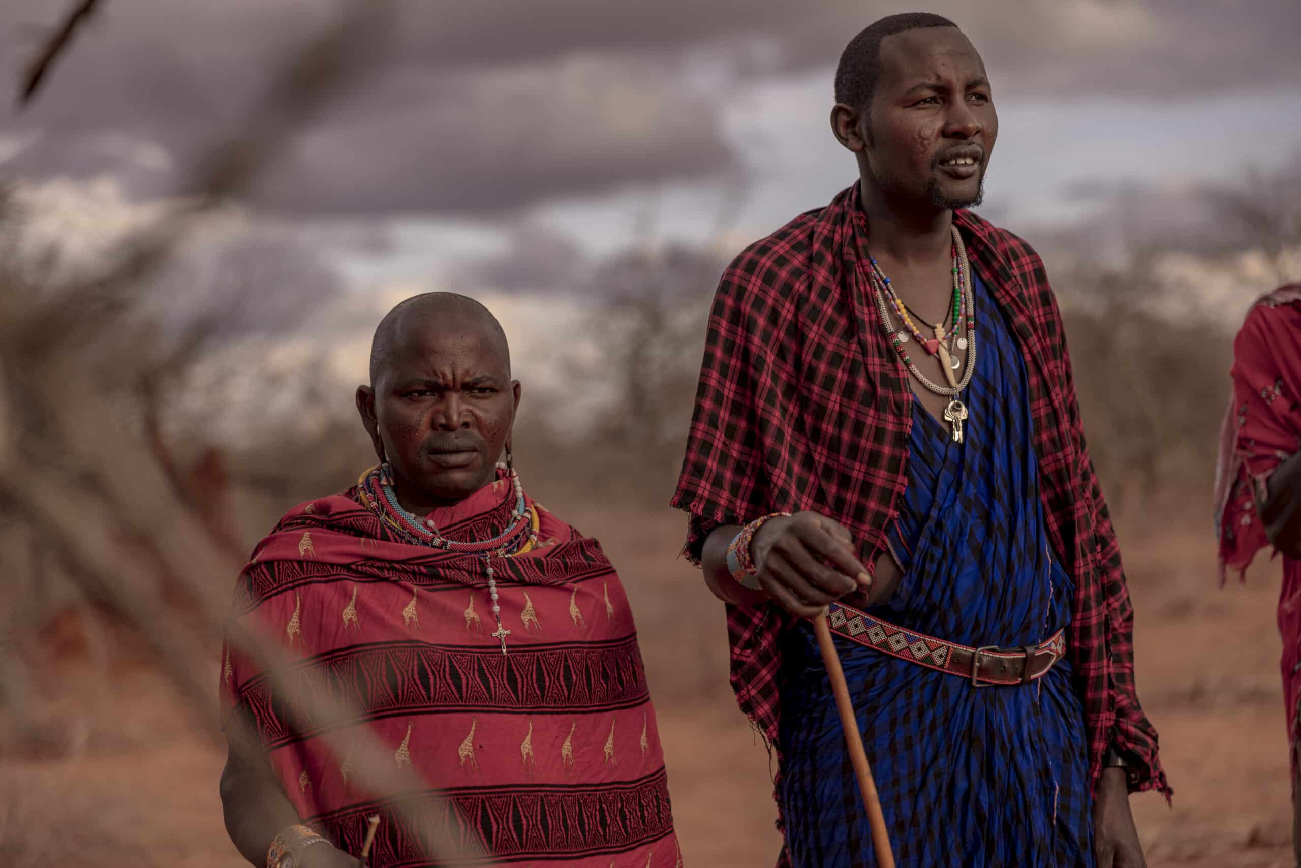 Zwei Massai-Männer in traditioneller Kleidung stehen an einem bewölkten Tag im Freien. Einer trägt einen roten und der andere einen blauen Shuka mit Perlenschmuck, blickt in die Ferne und verkörpert eine starke kulturelle Präsenz. © Fotografie Tomas Rodriguez
