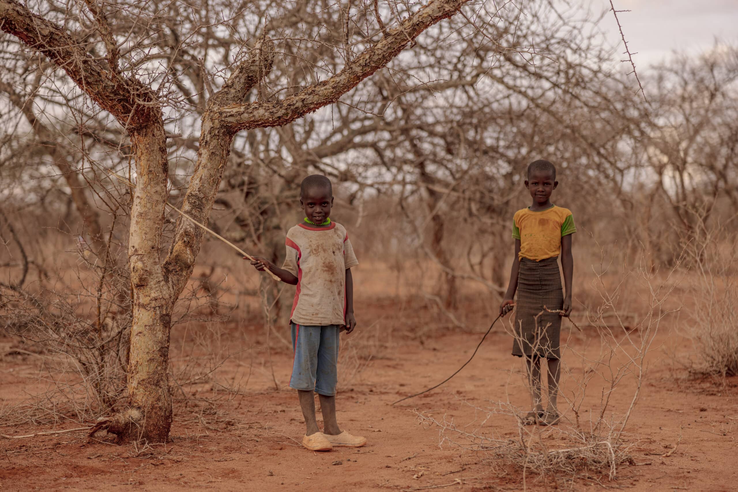 Zwei kleine Jungen stehen in einer trockenen, kargen Landschaft mit dünnen Bäumen und halten schmale Stöcke. Das Kind links trägt ein weiß-rotes Hemd, das rechte ein gelb-schwarzes Hemd. © Fotografie Tomas Rodriguez