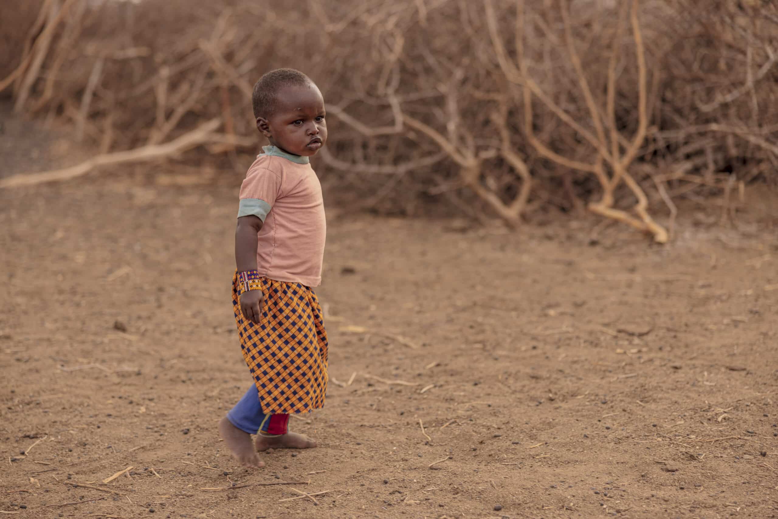 Ein kleines Kind steht in einer kargen Landschaft. Es trägt ein pfirsichfarbenes T-Shirt und einen gemusterten Rock. Im Hintergrund sind spärliche Vegetation und trockener Boden zu sehen. © Fotografie Tomas Rodriguez
