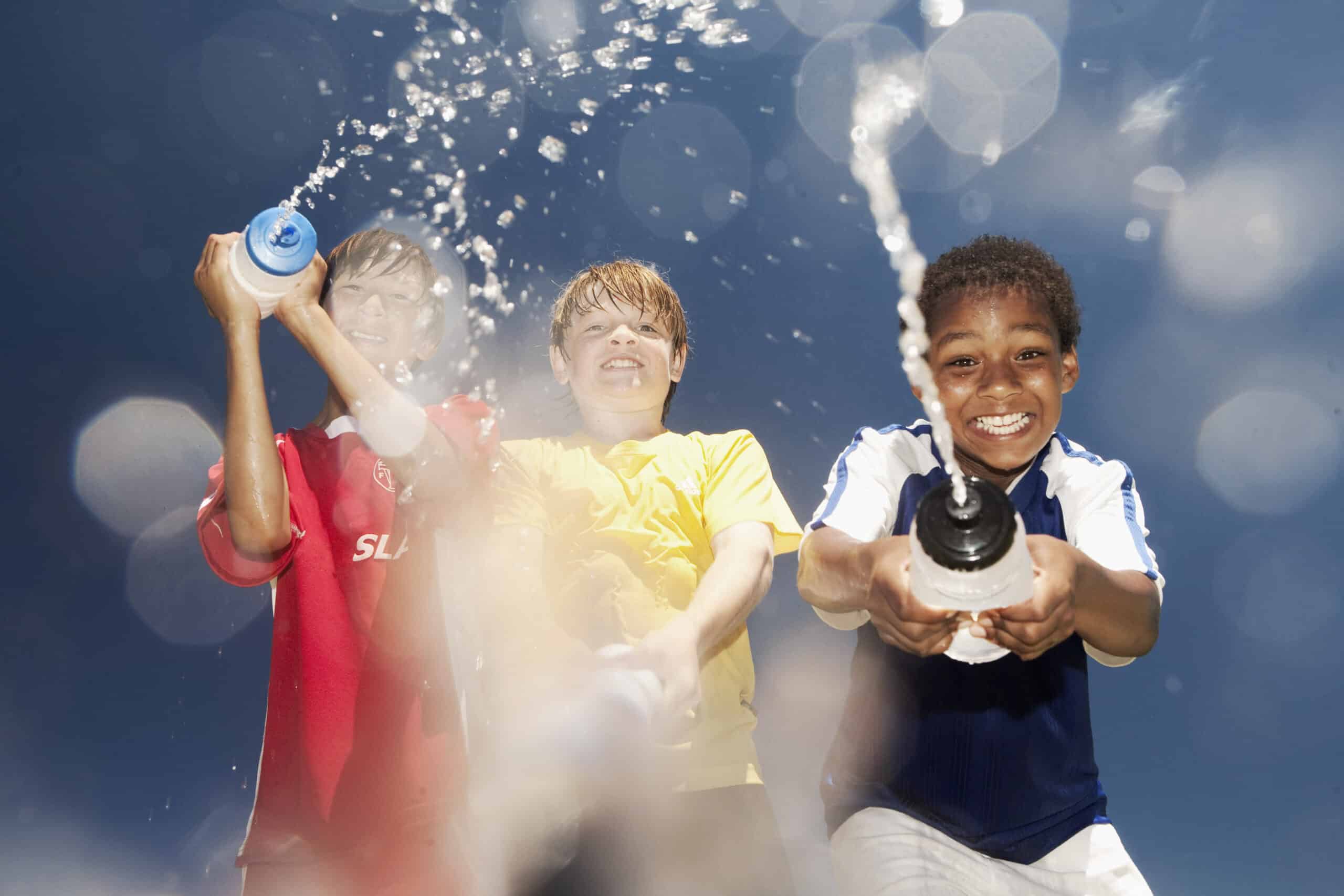 Drei Kinder spritzen fröhlich Wasser aus Flaschen unter einem strahlend blauen Himmel. Sie lächeln und tragen rote, gelbe und blaue Hemden. Wassertropfen und -spritzer sind in der Luft um sie herum eingefangen und unterstreichen den lustigen und energiegeladenen Moment. © Fotografie Tomas Rodriguez