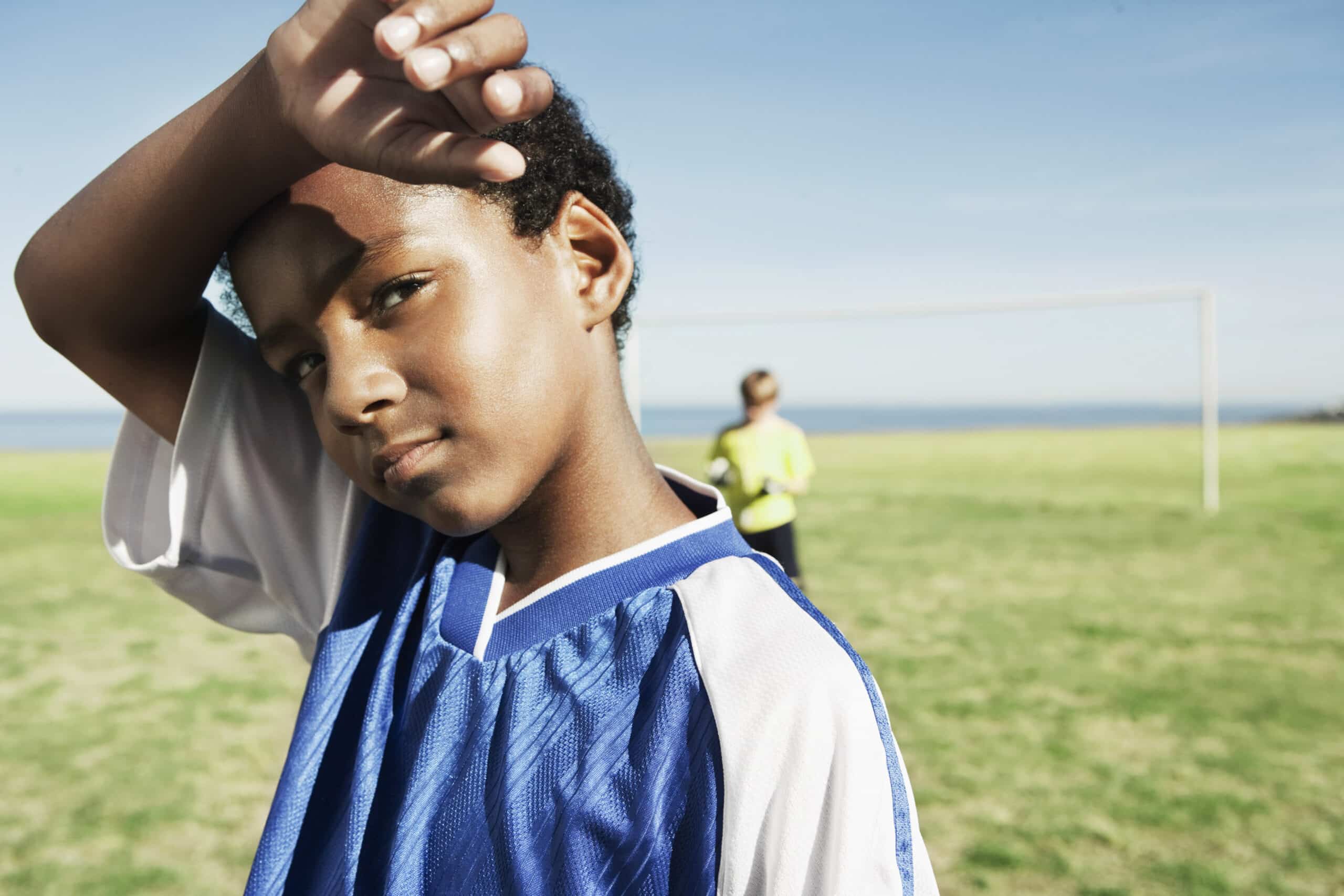 Ein kleiner Junge in einem blau-weißen Fußballtrikot wischt sich mit dem Arm die Stirn, während er auf einer Wiese steht. Im Hintergrund ist neben einem Fußballtor ein Kind in einem gelben Trikot zu sehen, und der Himmel ist klar und blau. © Fotografie Tomas Rodriguez
