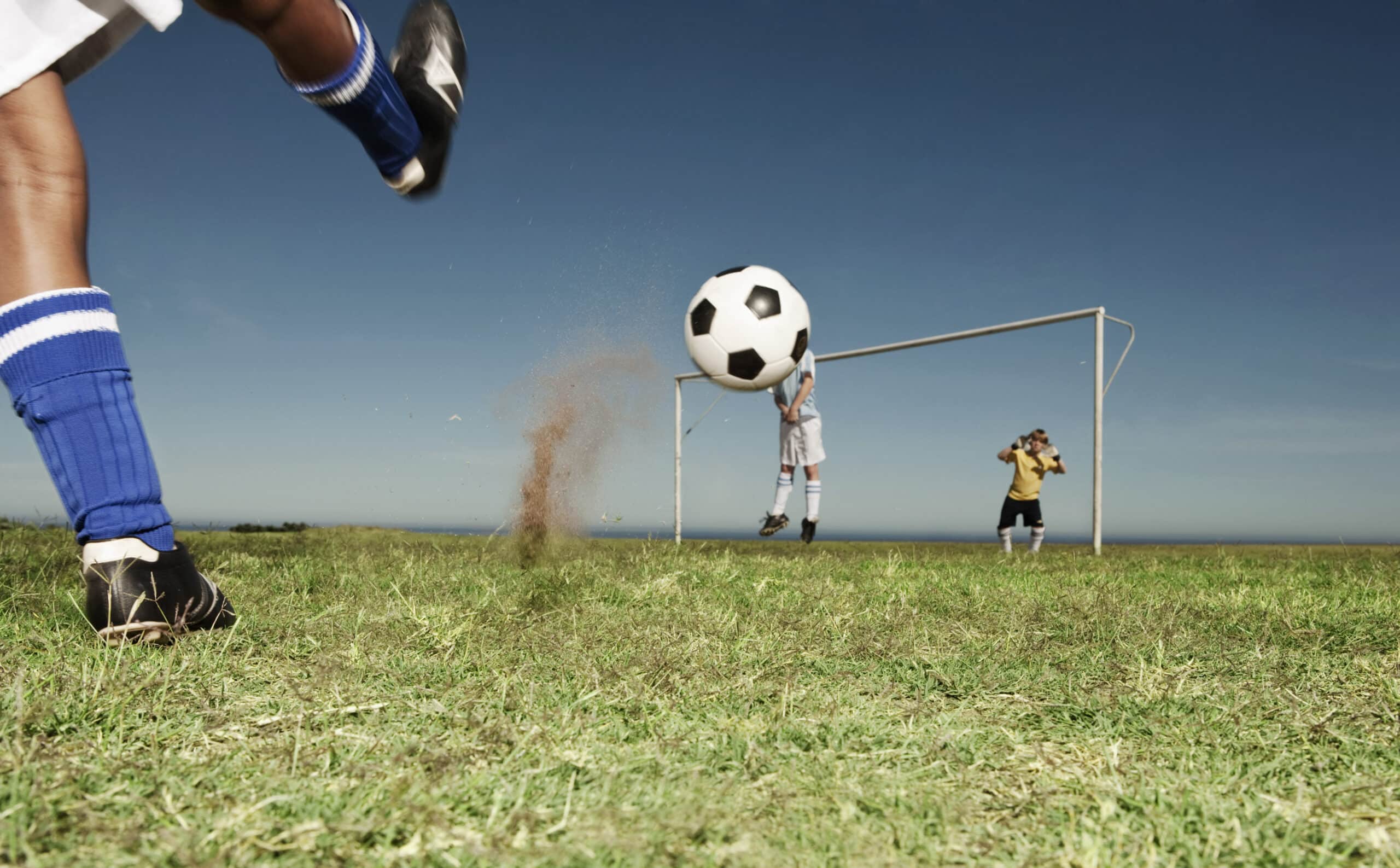 Ein Fußballspieler schießt einen Ball in Richtung Tor. Der Ball ist in der Luft und Staub fliegt neben dem Fuß des Schützen. Der Torwart in Gelb versucht, das Tor zu retten, während ein Spieler in Weiß daneben steht. Die Szene spielt sich auf einem Grasfeld unter einem klaren blauen Himmel ab. © Fotografie Tomas Rodriguez