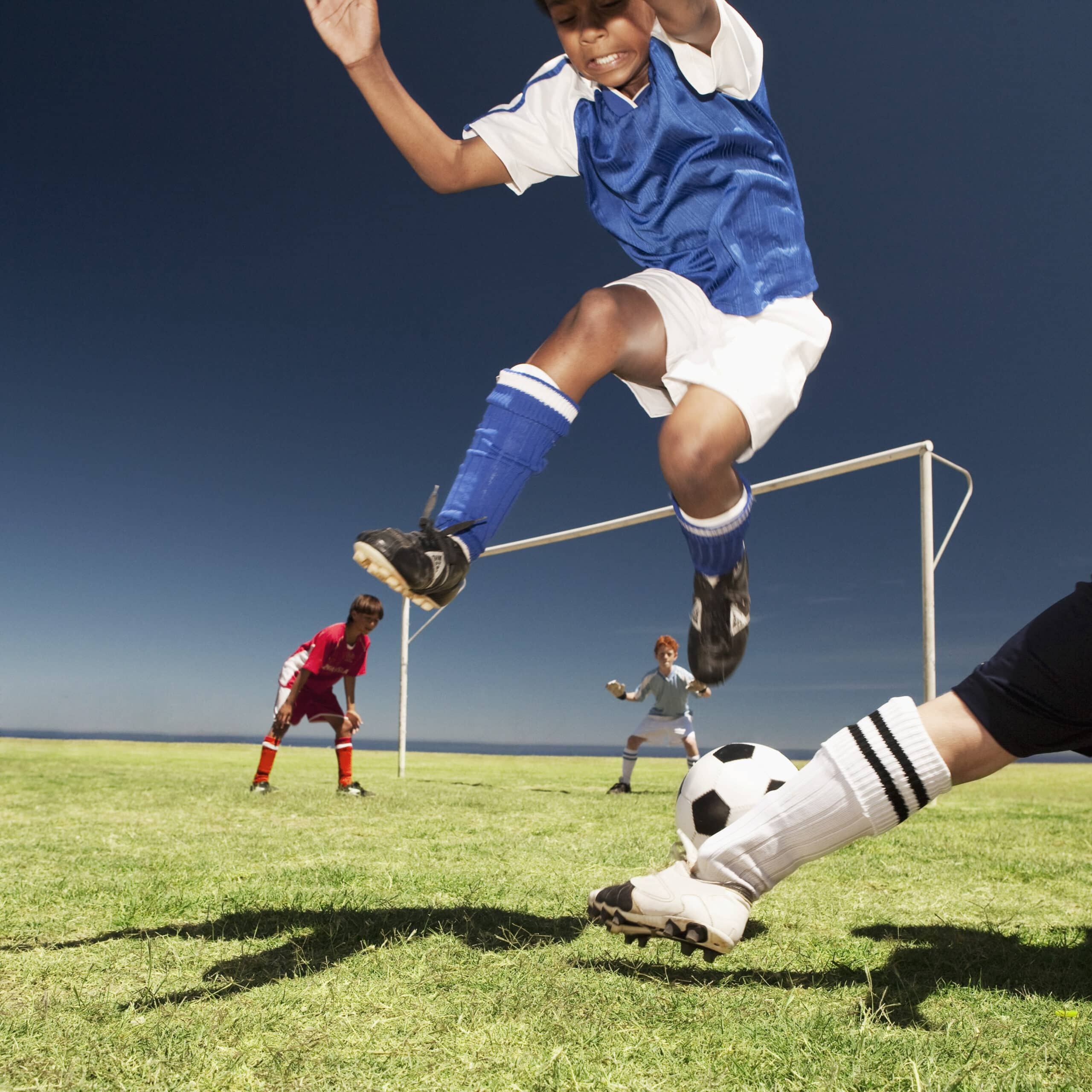 Ein junger Fußballspieler in blauem Trikot und weißen Shorts springt in die Luft und versucht, einen Fußball auf einem Rasenplatz zu kicken. Ein weiterer Spieler in weißen Socken und schwarzen Stollenschuhen greift ebenfalls nach dem Ball. Im Hintergrund sind zwei weitere Spieler und ein Torpfosten zu sehen. © Fotografie Tomas Rodriguez