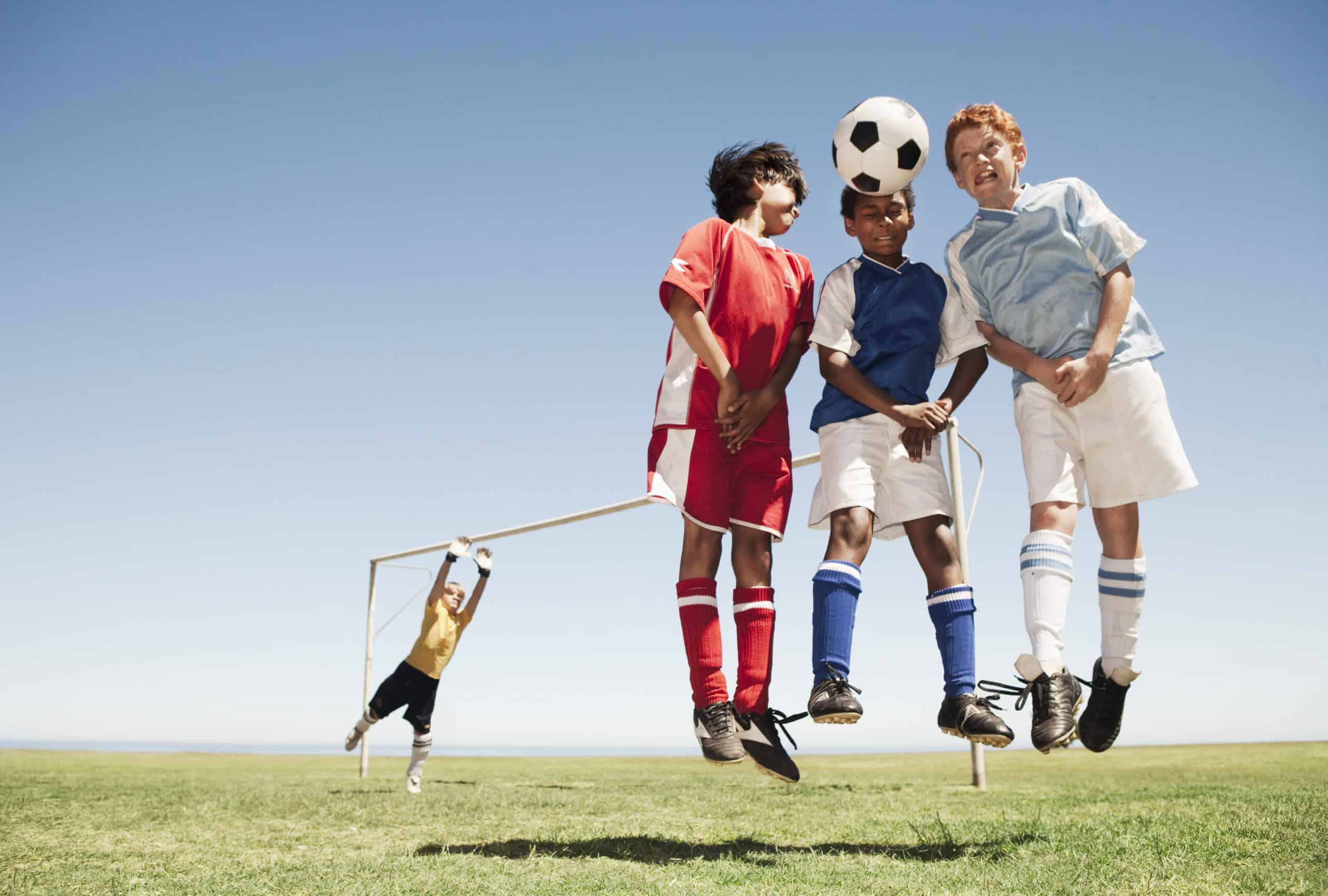 Vier Jungen in Fußballtrikots springen, um einen Fußball zu köpfen. Sie stehen auf einem Rasenplatz unter einem klaren blauen Himmel. Hinter ihnen streckt sich ein Torwart im gelben Trikot aus und versucht, einen Ball am Torpfosten abzuwehren. © Fotografie Tomas Rodriguez