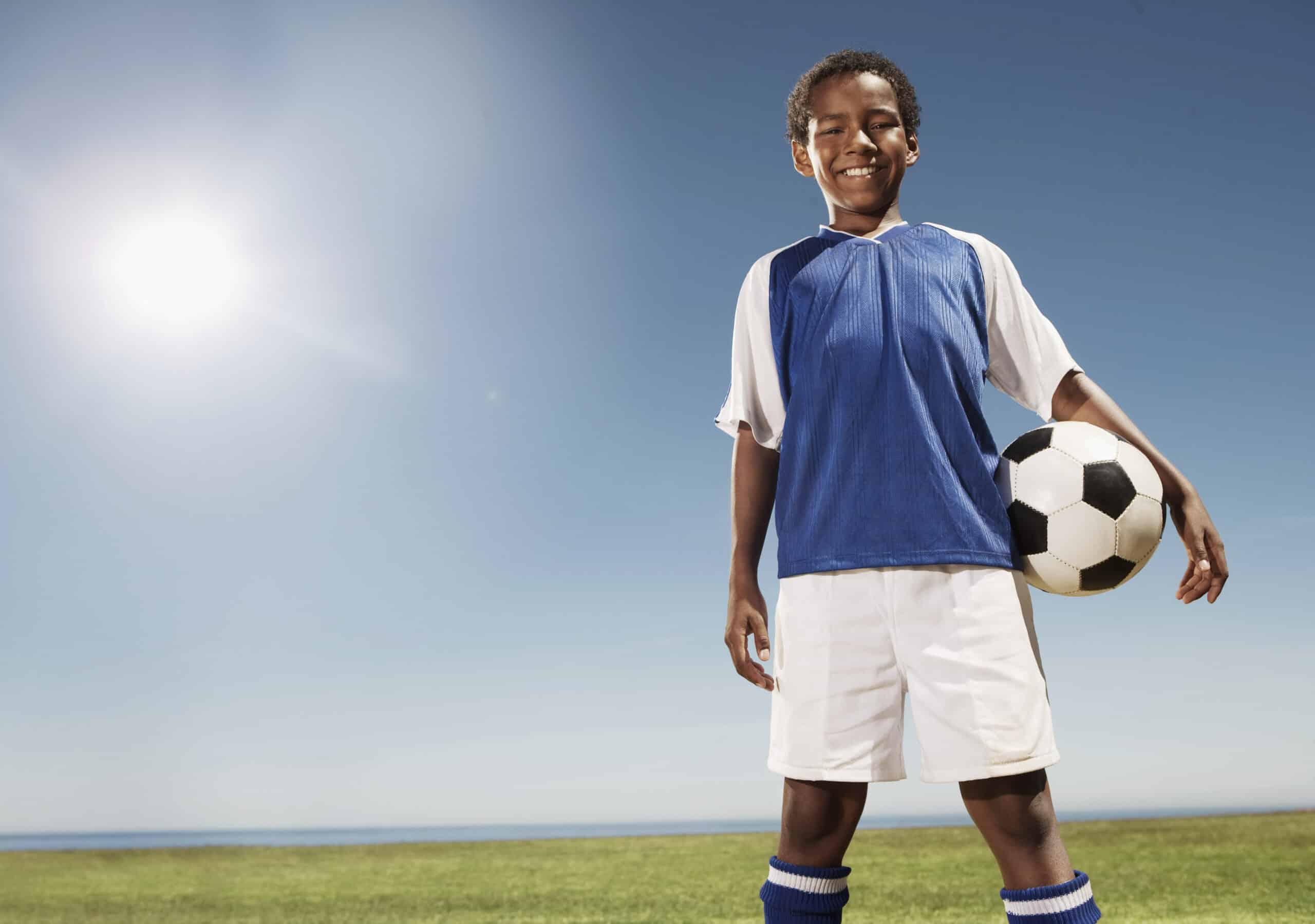 Ein lächelndes Kind in einer blau-weißen Fußballuniform steht auf einem Rasenplatz und hält einen Fußball unter dem Arm. Die Sonne scheint hell am klaren blauen Himmel darüber. © Fotografie Tomas Rodriguez