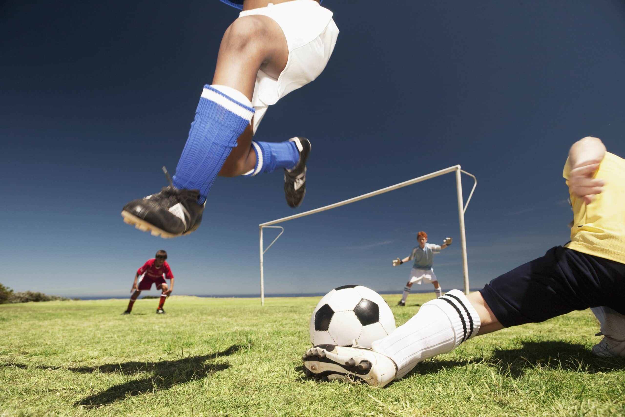 Nahaufnahme eines Fußballspiels auf einem Rasenplatz. Ein Spieler in blauen Shorts und blauen Socken springt in der Luft über einen Spieler in Gelb, der den Fußball angreift. Im Hintergrund stehen zwei weitere Spieler in der Nähe des Torpfostens, einer in Rot und einer in Grau. © Fotografie Tomas Rodriguez