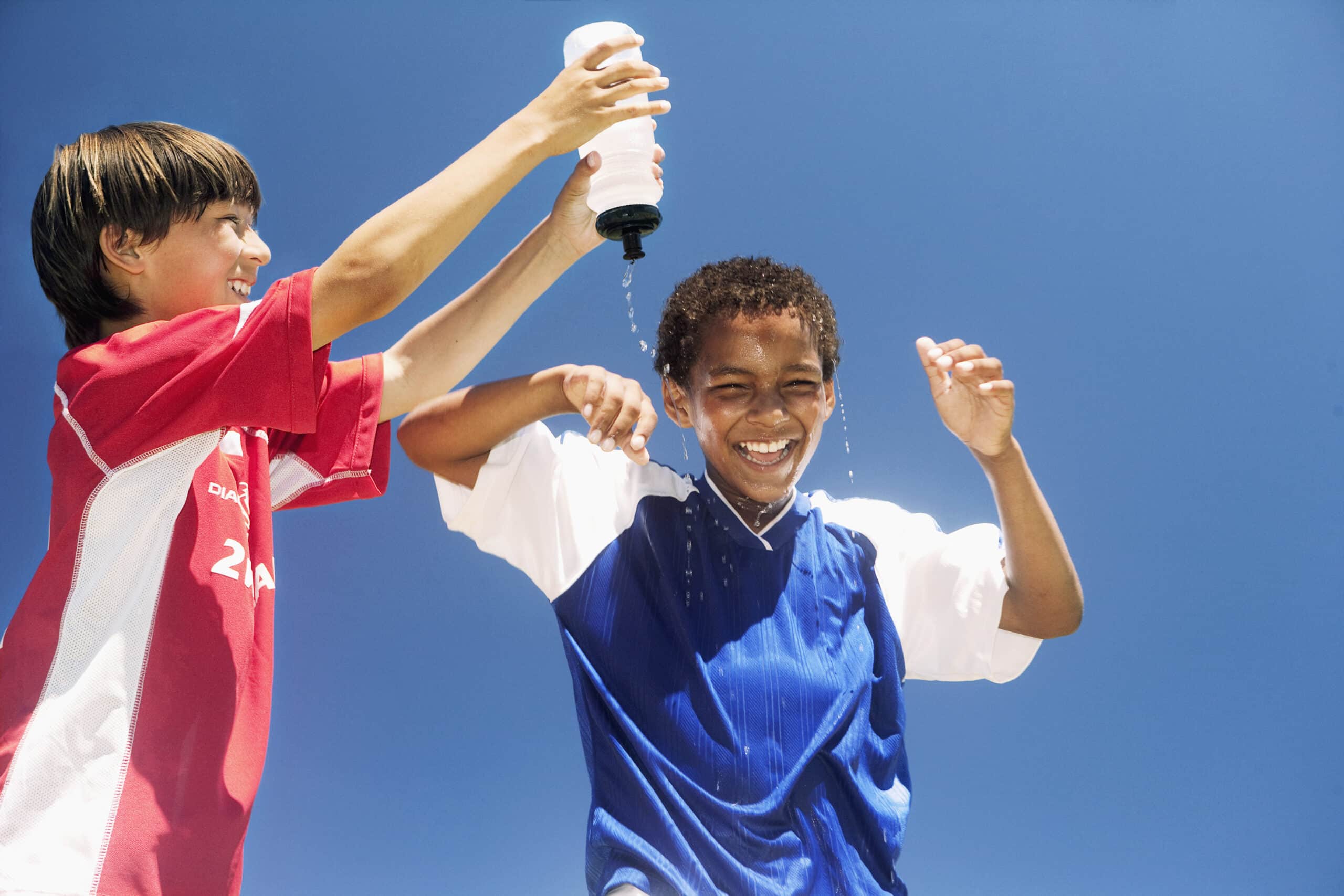 Zwei Jungen in Fußballtrikots haben Spaß unter einem klaren blauen Himmel. Ein Junge in Rot gießt Wasser aus einer Flasche über den Kopf des anderen Jungen in Blau, der lacht und den Moment genießt. © Fotografie Tomas Rodriguez