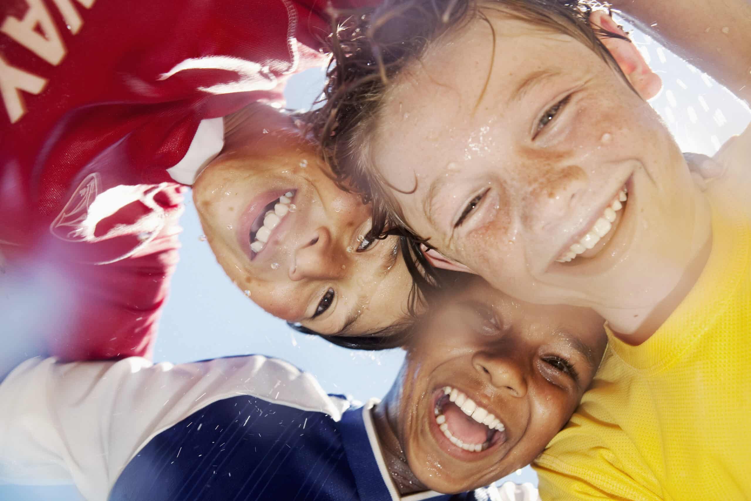 Drei Kinder in Sporttrikots lächeln und blicken in die Kamera. Sie wirken glücklich und verschwitzt, wahrscheinlich nach einem Spiel. Im Hintergrund ist ein klarer blauer Himmel zu sehen. Das Foto fängt einen Moment der Freude und Freundschaft ein. © Fotografie Tomas Rodriguez