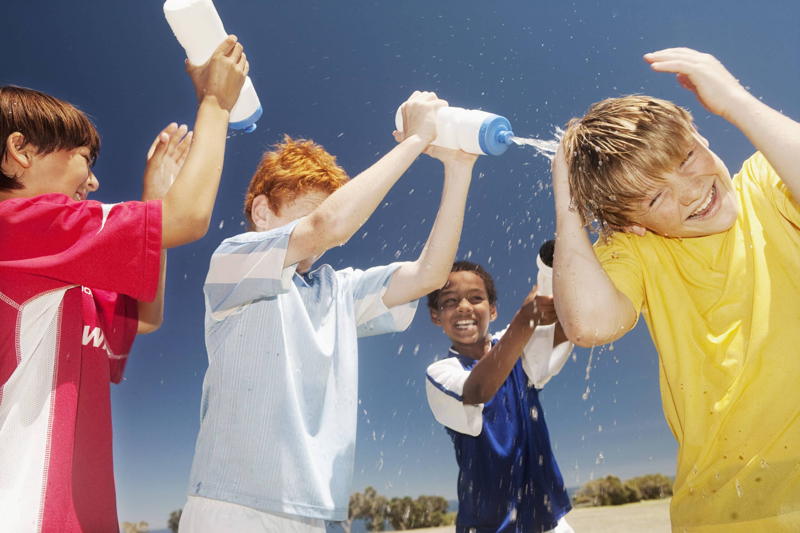 Vier Jungen lachen und spielen und gießen sich an einem sonnigen Tag gegenseitig Wasser aus Flaschen über den Kopf. Ein Junge im roten Hemd klatscht, ein Junge im blauen Hemd gießt einem anderen Wasser über den Kopf und ein Junge im gelben Hemd grinst, als Wasser auf ihn spritzt. Der Himmel ist klar und blau. © Fotografie Tomas Rodriguez