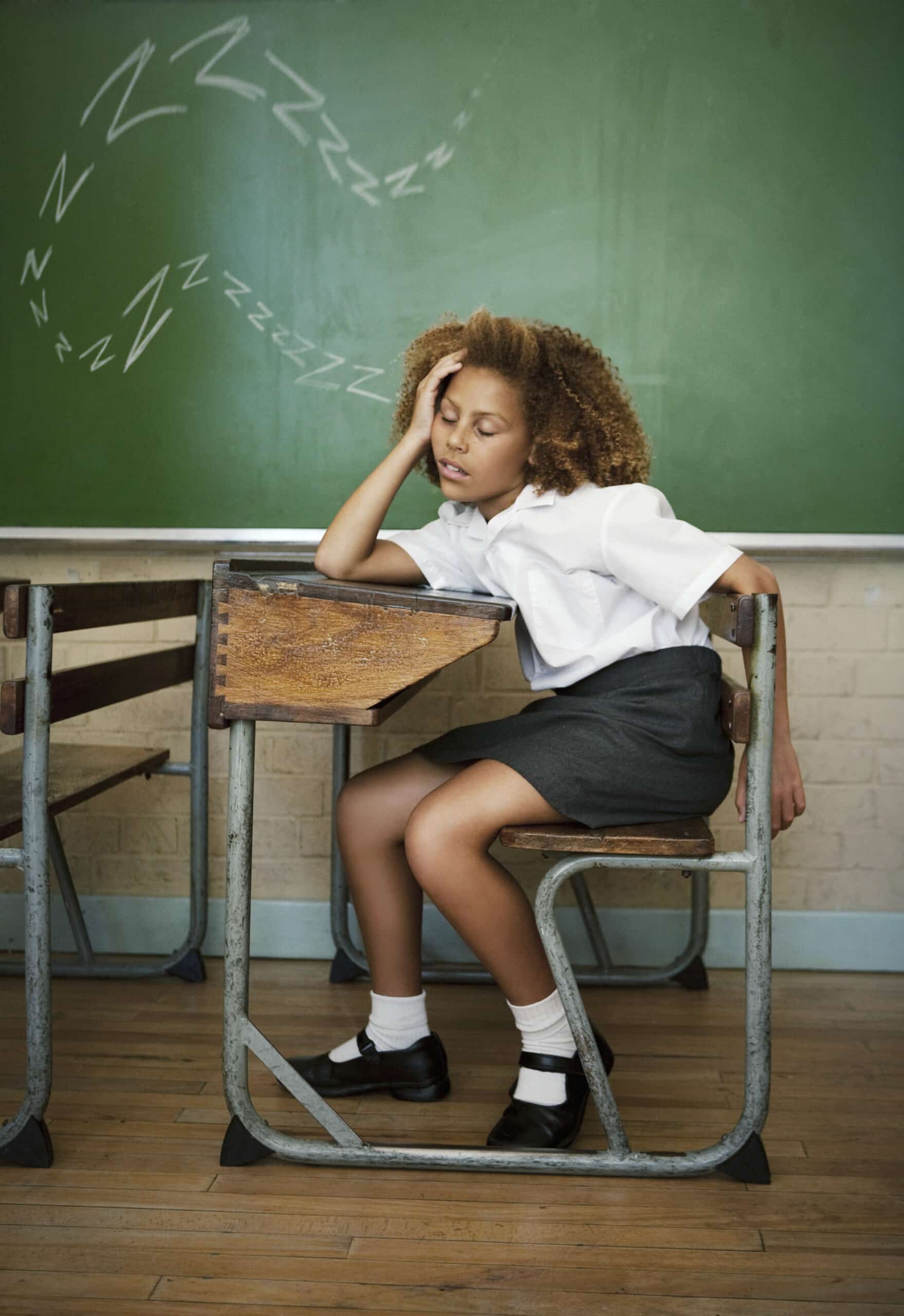 Ein junger Schüler in Schuluniform sitzt zusammengesunken über einem Holztisch und scheint zu schlafen. Das Klassenzimmer hat eine grüne Tafel und einen Holzboden. Ein gezeichnetes „Zzz“-Symbol über dem Kopf des Schülers zeigt an, dass er schläft. © Fotografie Tomas Rodriguez