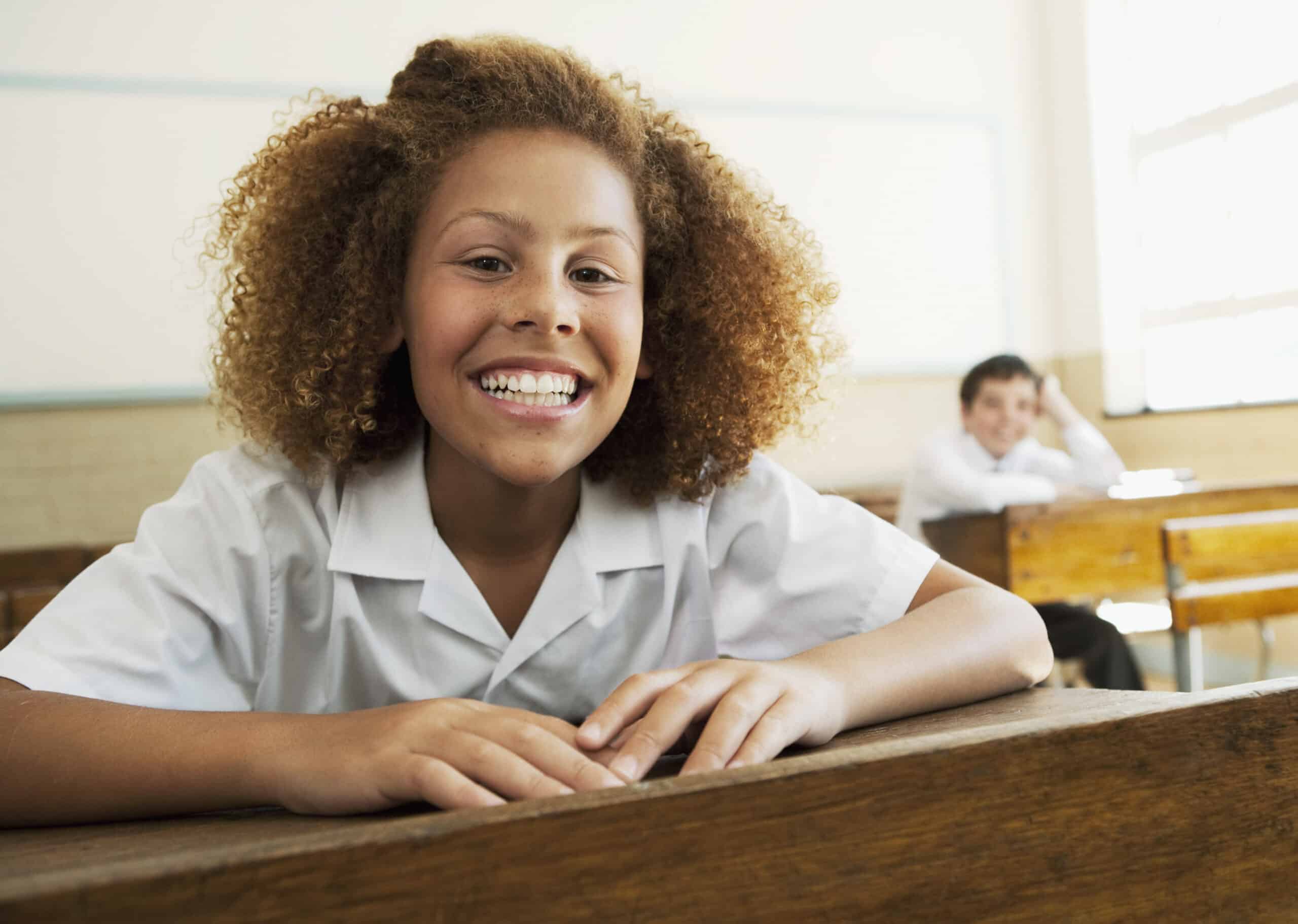Ein junger Student in weißem Hemd lehnt sich an einen Holztisch in einem Klassenzimmer und lächelt strahlend. Im Hintergrund ist ein weiterer Student zu sehen, der an einem Tisch sitzt und in die Kamera blickt. Das Klassenzimmer hat große Fenster und ein helles Interieur. © Fotografie Tomas Rodriguez