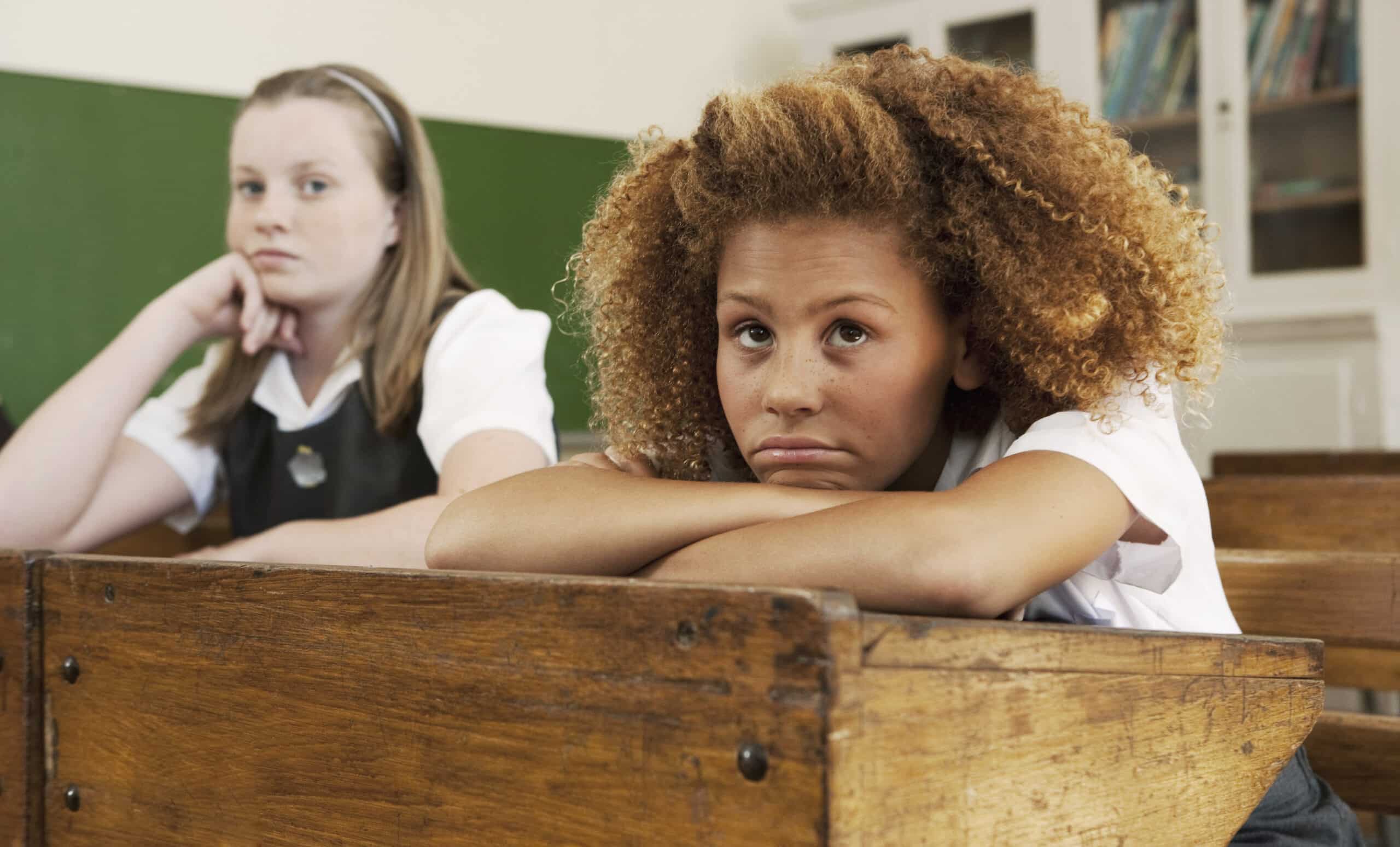 Zwei Schülerinnen in einem Klassenzimmer. Das Mädchen im Vordergrund hat lockiges Haar und sieht gelangweilt aus. Es stützt sein Kinn auf die Hände auf dem Tisch. Das Mädchen im Hintergrund trägt ein Stirnband und blickt nach vorne. Es sitzt an einem Holztisch. Beide tragen Schuluniformen. © Fotografie Tomas Rodriguez