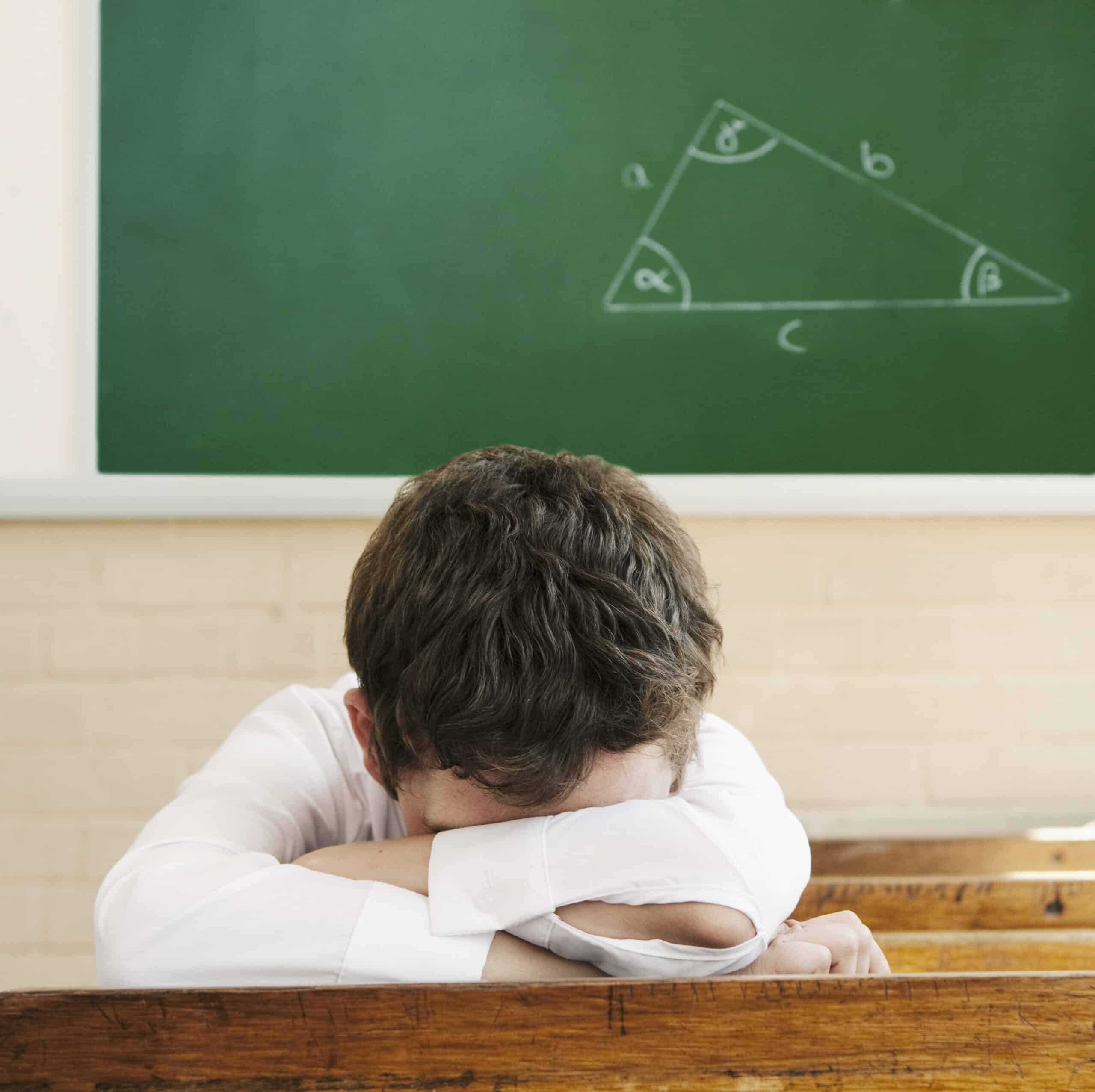 Ein Student mit braunem Haar und weißem Hemd sitzt an einem Holztisch vor einer Tafel. Der Kopf des Studenten ruht auf seinen verschränkten Armen und blickt nach unten. An der Tafel ist ein Diagramm eines rechtwinkligen Dreiecks mit beschrifteten Seiten und Winkeln angebracht. © Fotografie Tomas Rodriguez