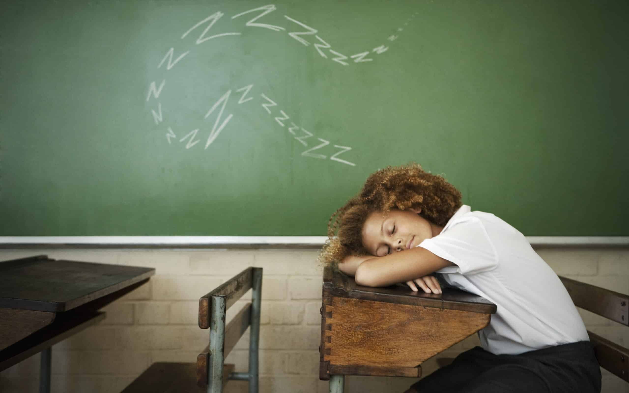 Ein Schüler mit lockigem Haar und weißem Hemd schläft an seinem Schreibtisch in einem Klassenzimmer. Der Schreibtisch besteht aus Holz und Metall und überall stehen leere Schreibtische. Auf der grünen Tafel hinter ihnen sind eine Reihe von „Zzz“-Symbolen gezeichnet, die anzeigen, dass sie schlafen. © Fotografie Tomas Rodriguez
