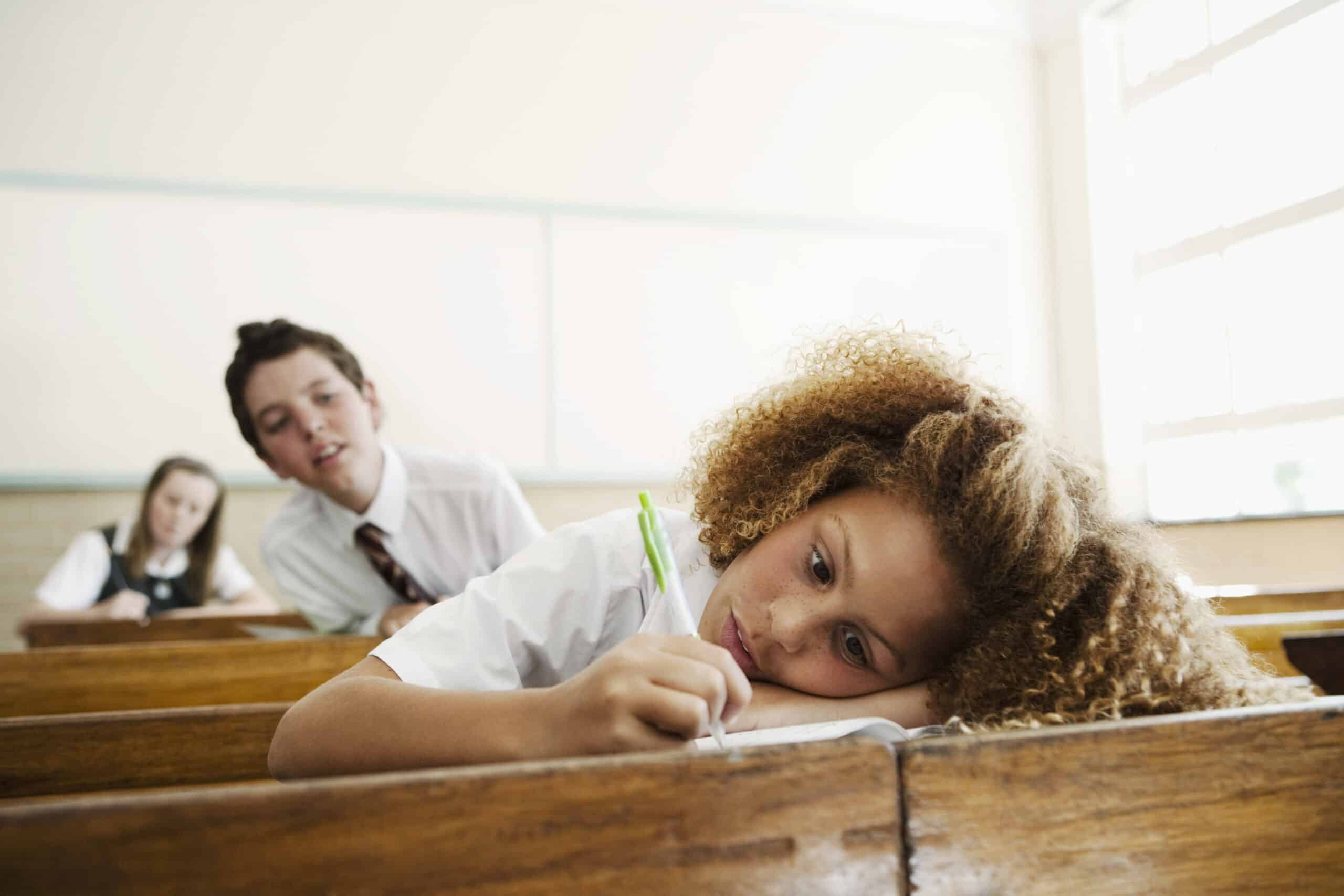 Eine junge Studentin mit lockigem Haar lehnt an ihrem Schreibtisch und schreibt mit einem grünen Stift in ein Notizbuch. Hinter ihr blickt ein weiterer Student mit Krawatte in ihre Richtung. Ein dritter Student sitzt weiter hinten im Klassenzimmer am Fenster. © Fotografie Tomas Rodriguez