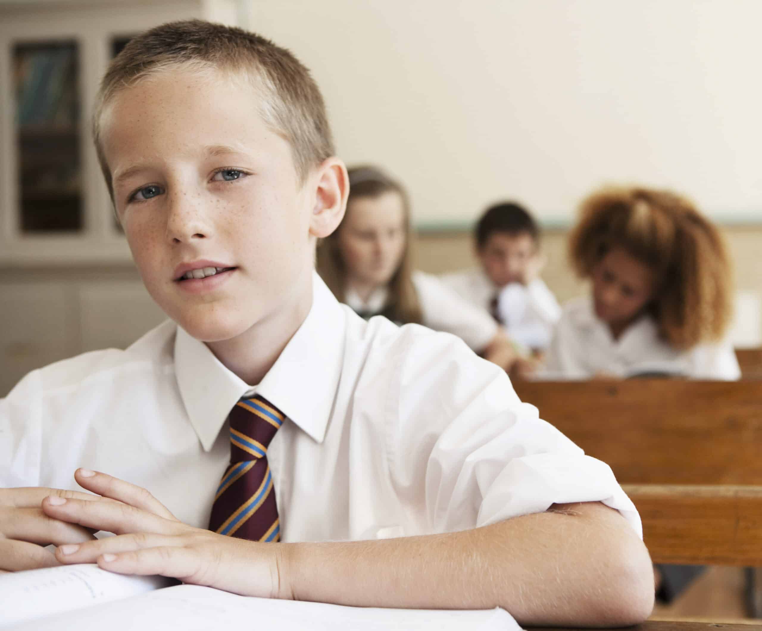 Ein kleiner Junge mit kurzen Haaren sitzt in einem Klassenzimmer an einem Schreibtisch, trägt ein weißes Hemd und eine gestreifte Krawatte und blickt in die Kamera. Im Hintergrund sitzen andere Schüler und sind mit ihren eigenen Aktivitäten beschäftigt. Die Unterrichtsumgebung lässt an eine Schulumgebung denken. © Fotografie Tomas Rodriguez