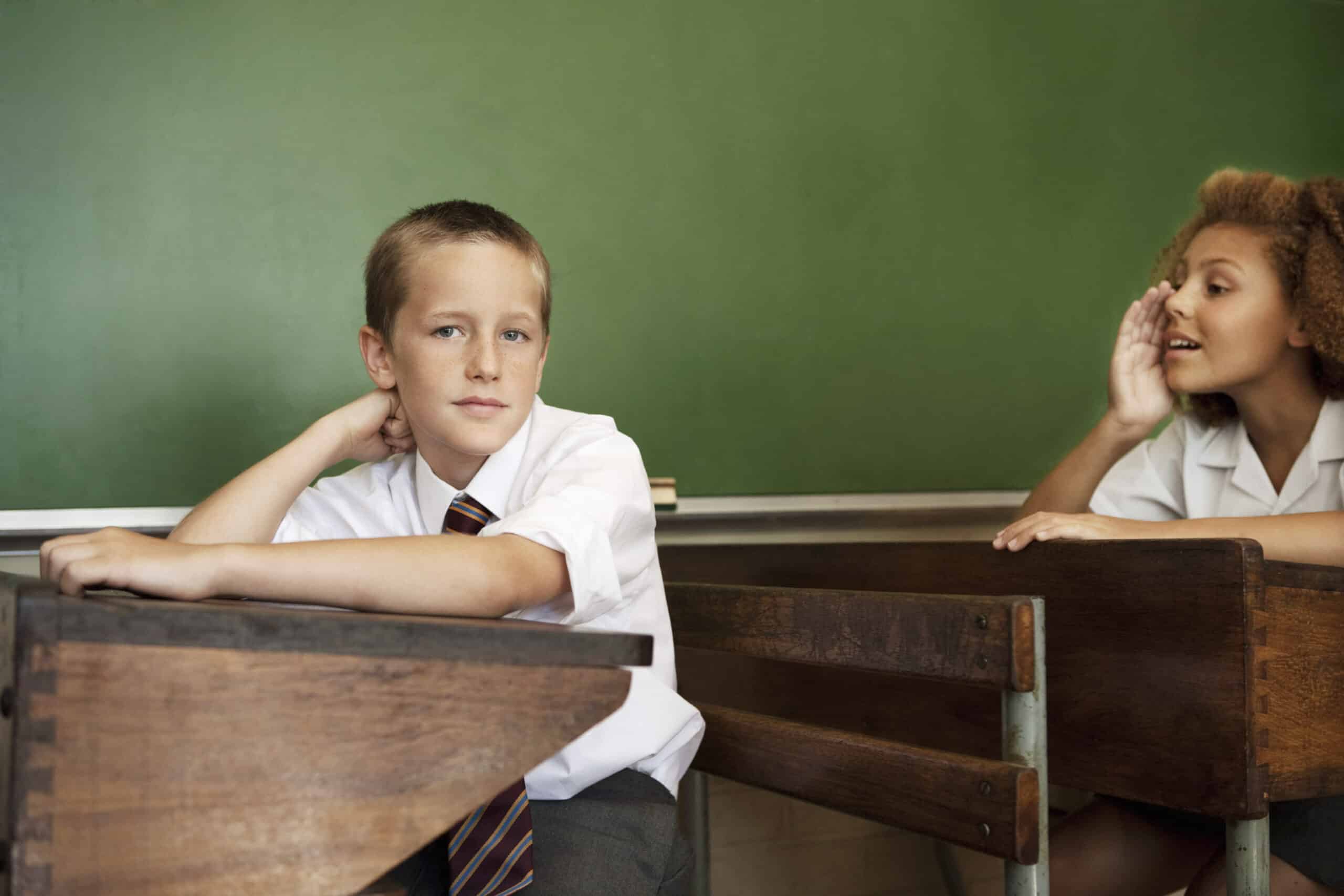 Ein Junge in Schuluniform sitzt an einem Holzpult in einem Klassenzimmer und blickt nachdenklich in die Kamera. Ein Mädchen in Schuluniform sitzt an einem nahegelegenen Pult, beugt sich nach vorne und flüstert oder spricht in seine Richtung. Im Hintergrund ist eine grüne Tafel zu sehen. © Fotografie Tomas Rodriguez